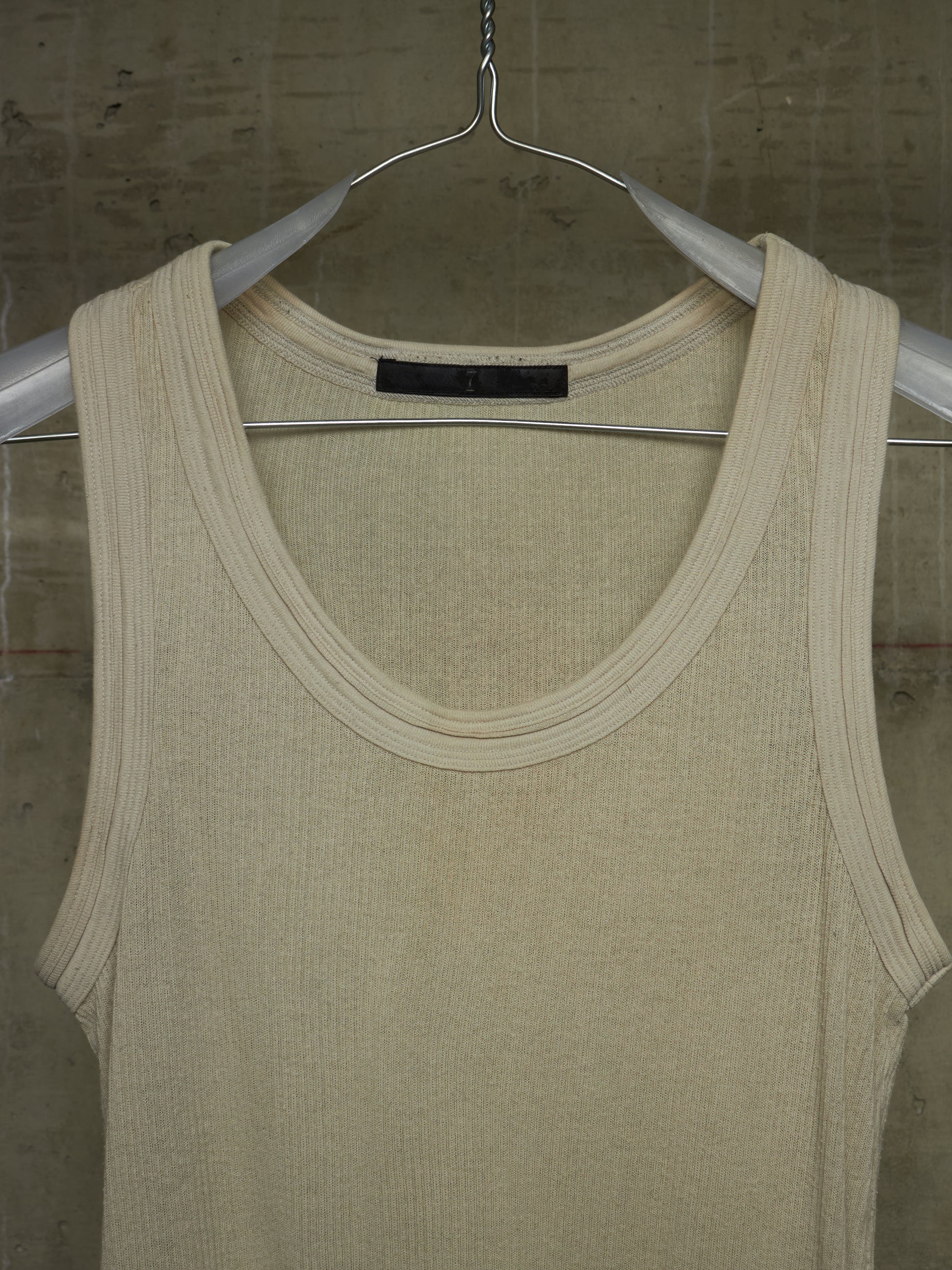 A cream-colored ribbed knit tank top hanging on a silver metal hanger against a textured concrete wall.