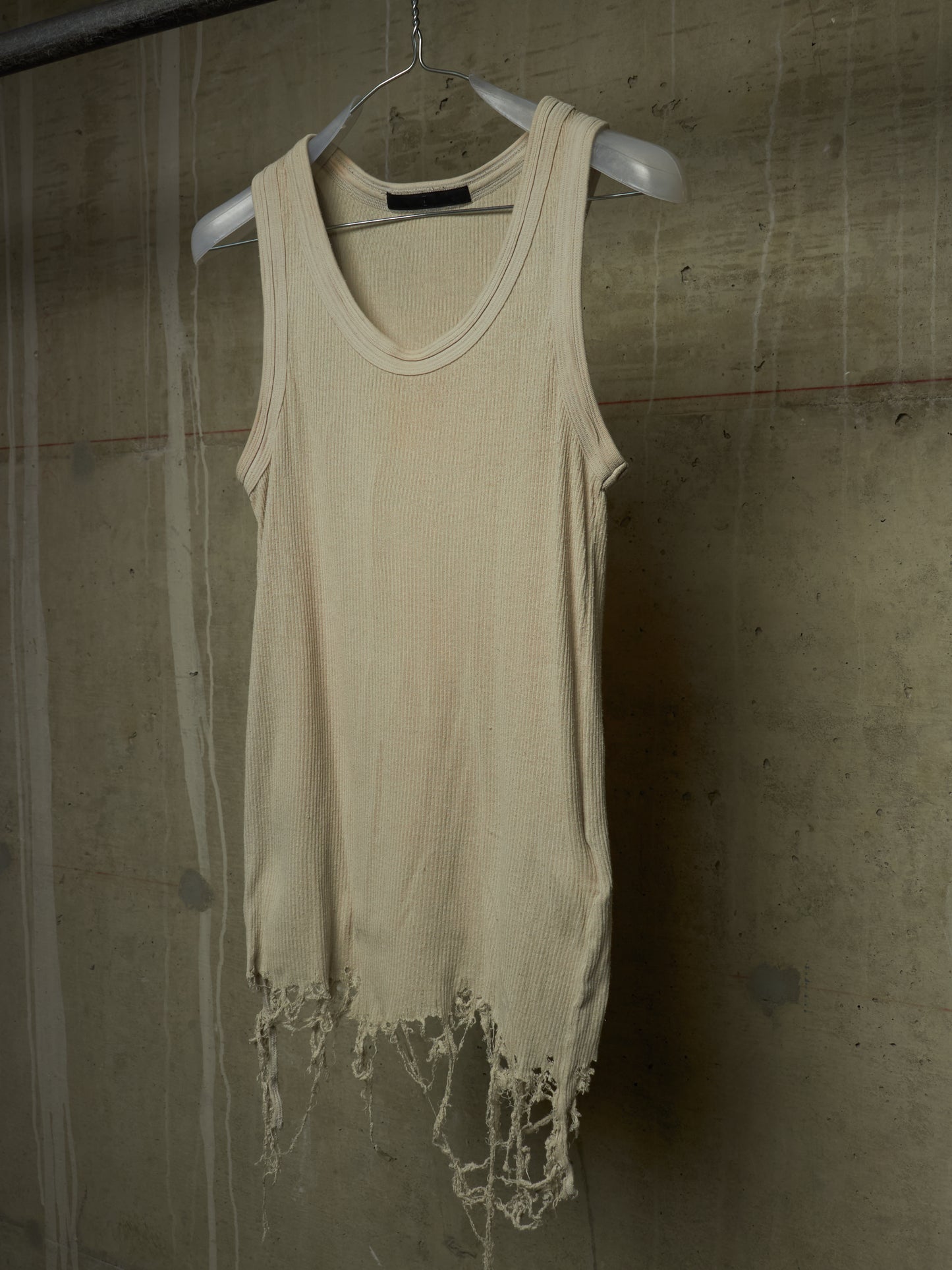 A cream-colored, sleeveless distressed tank top with frayed edges, hanging on a white plastic hanger against a textured concrete wall.