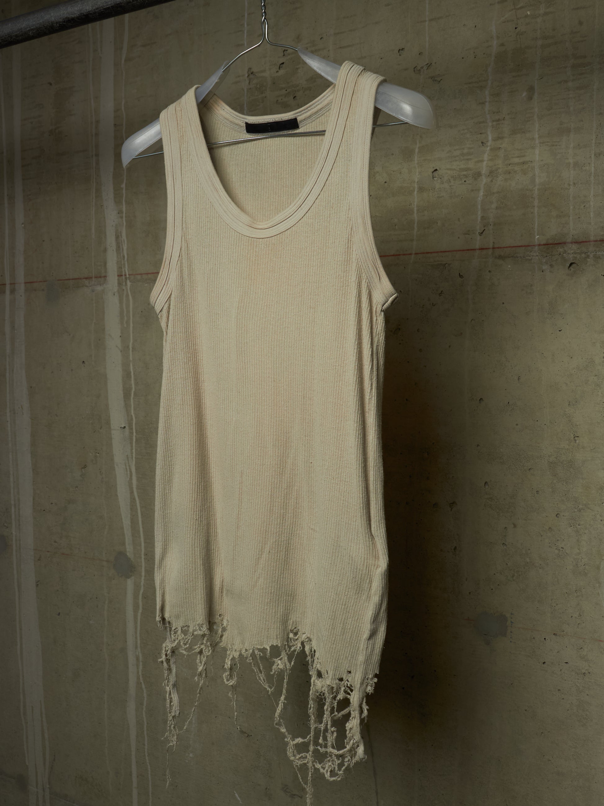 A cream-colored, sleeveless distressed tank top with frayed edges, hanging on a white plastic hanger against a textured concrete wall.