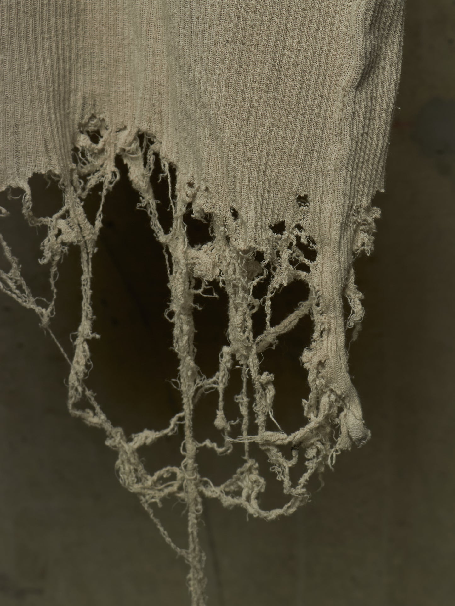 A close-up of a distressed, tattered ribbed knit fabric in a muted beige-gray color, hanging against a dark gray background.