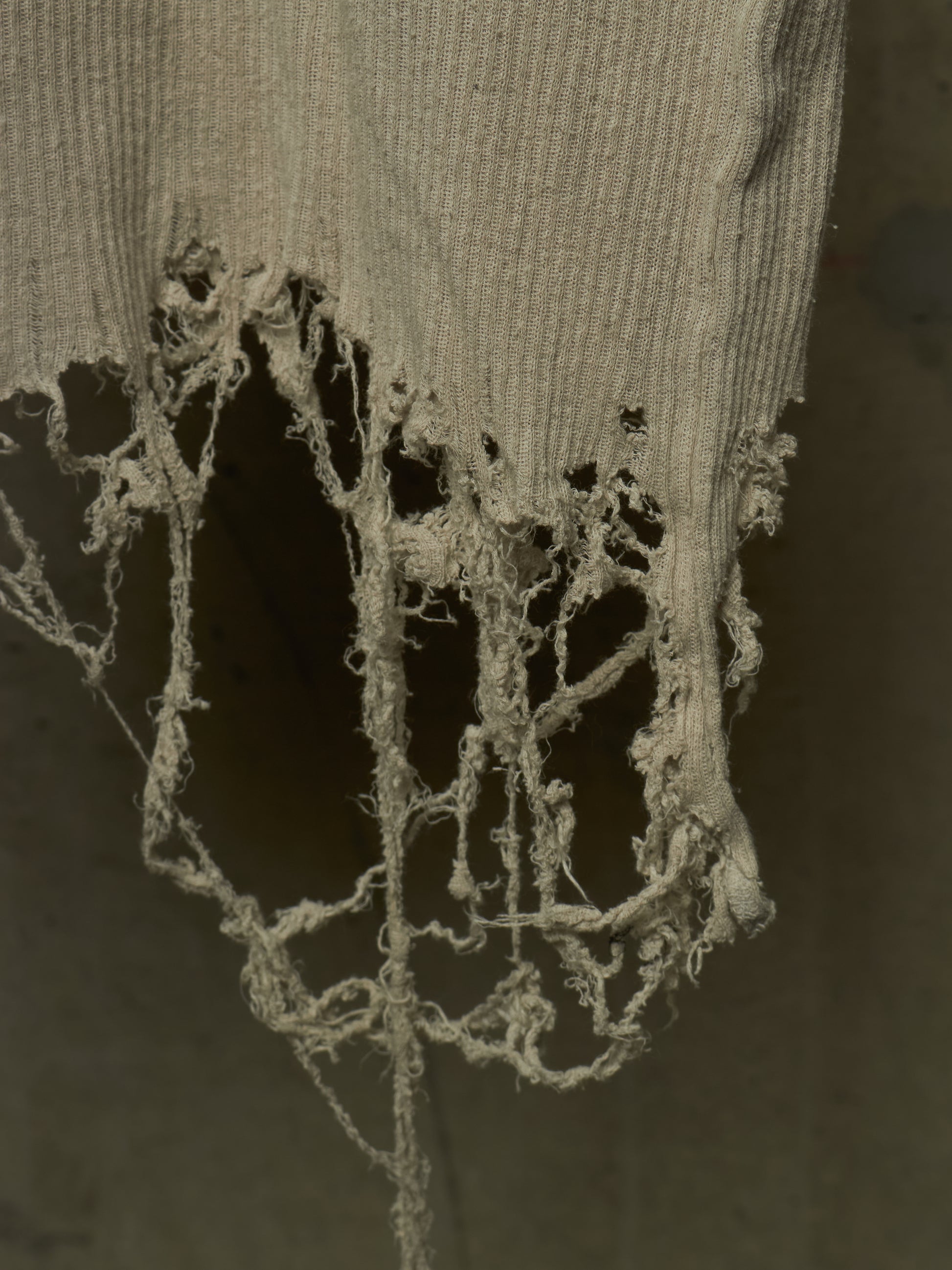 A close-up of a distressed, tattered ribbed knit fabric in a muted beige-gray color, hanging against a dark gray background.