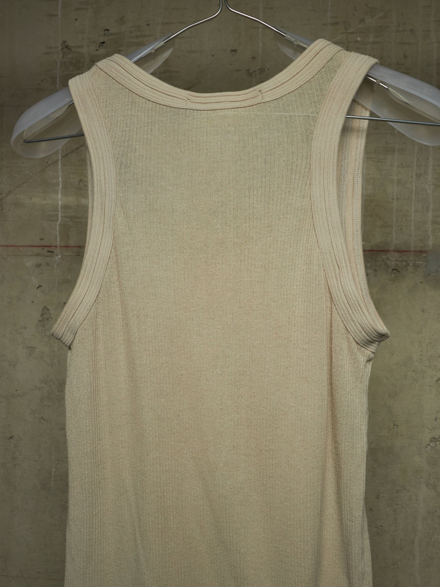 A cream-colored ribbed knit tank top hanging on a clear plastic hanger against a textured concrete wall.
