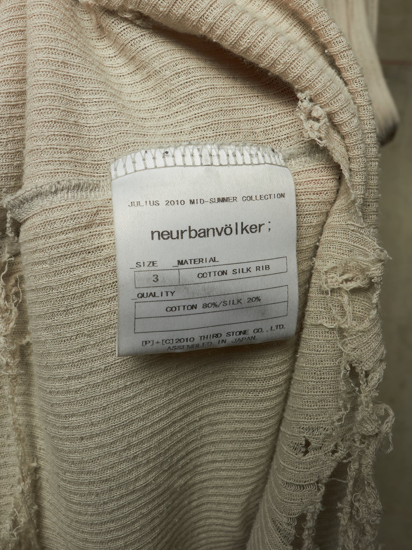 Close-up of a beige ribbed knit garment with a white sewn-in label reading 'neurbanvölker' and product details, showing frayed textured fabric around the collar area.