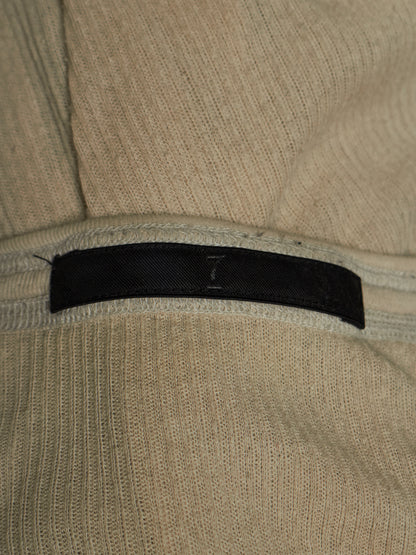 Close-up of a beige ribbed knit garment with a black fabric label sewn horizontally across the chest.