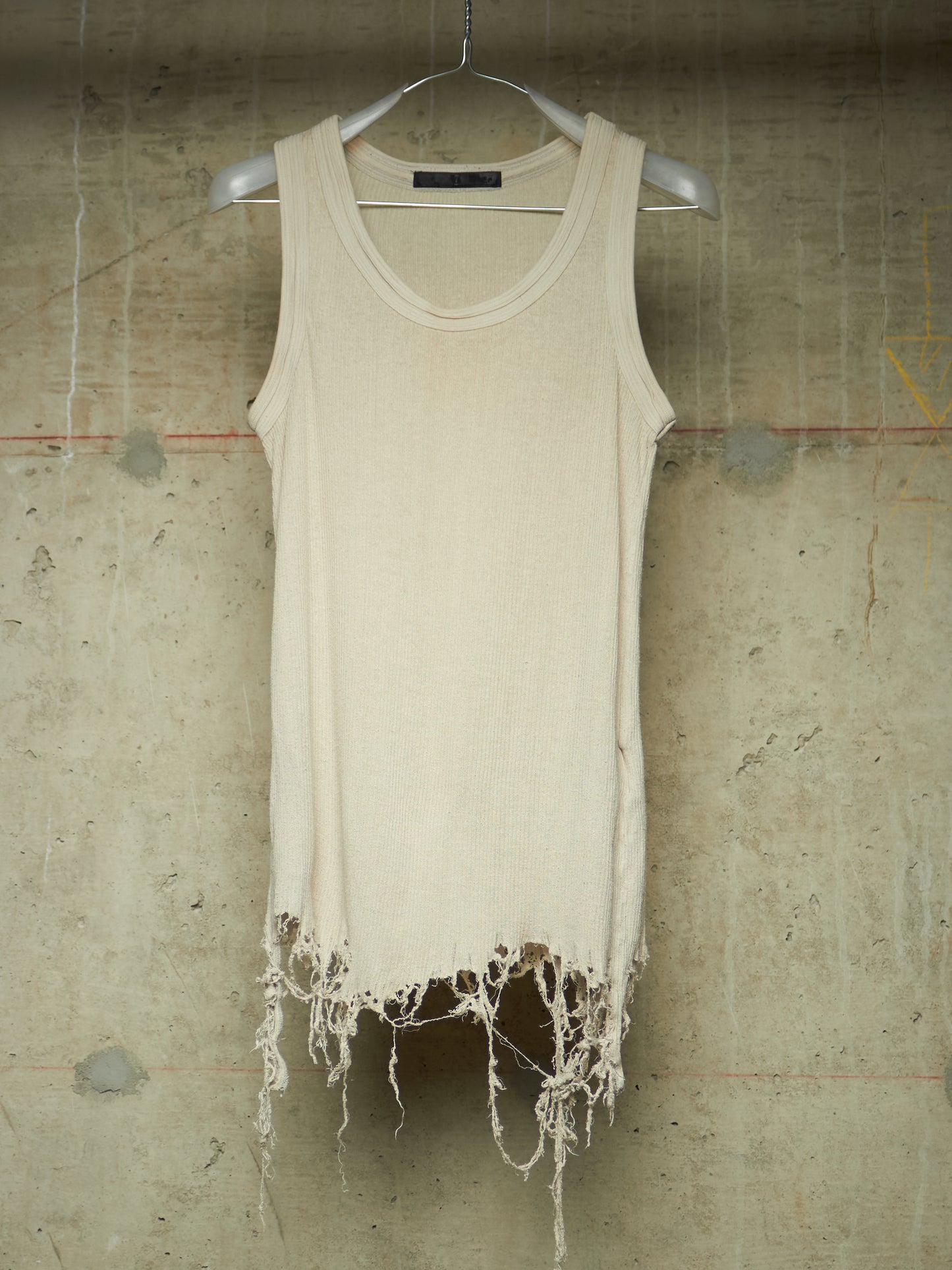 A cream-colored sleeveless knit top with a raw, frayed hem, hanging on a white hanger against a textured concrete wall.