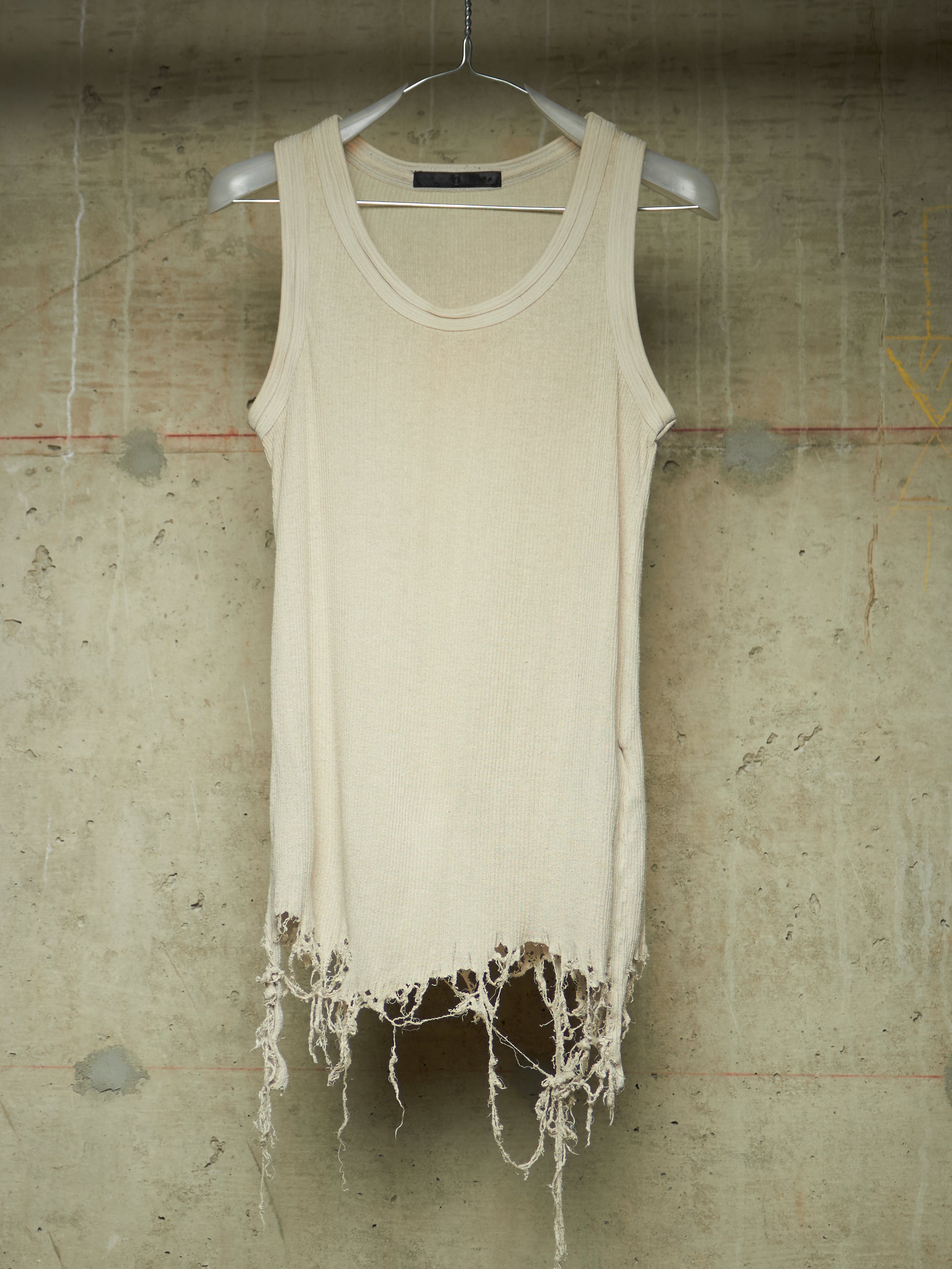 A cream-colored sleeveless knit top with a raw, frayed hem, hanging on a white hanger against a textured concrete wall.