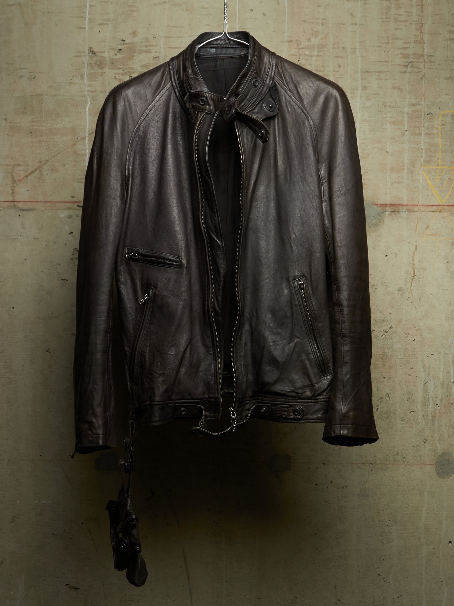 A dark brown leather motorcycle jacket hanging on a metal hanger against a textured concrete wall.