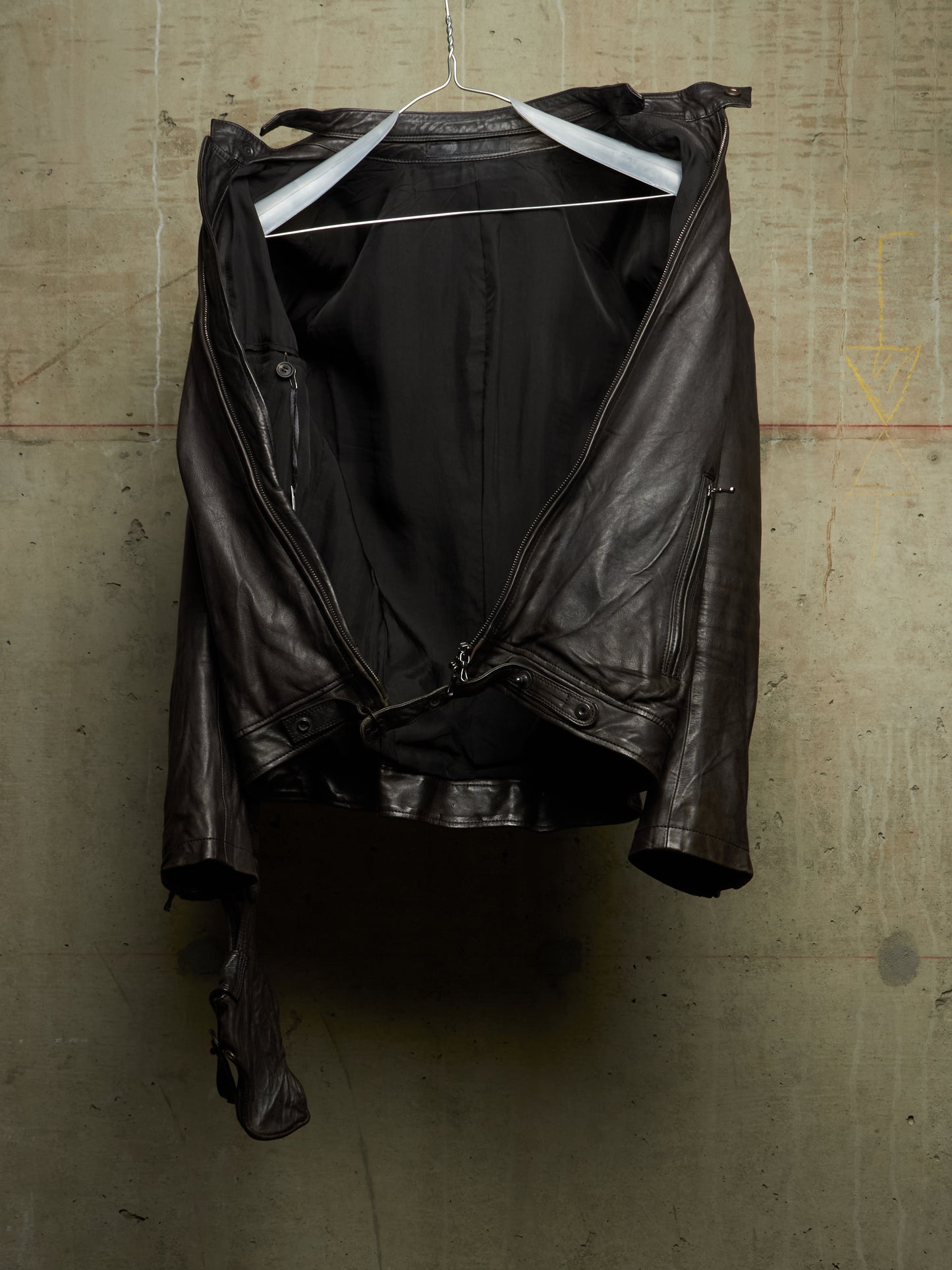 A dark brown leather jacket with a black lining, hanging on a white wire hanger against a textured concrete wall.