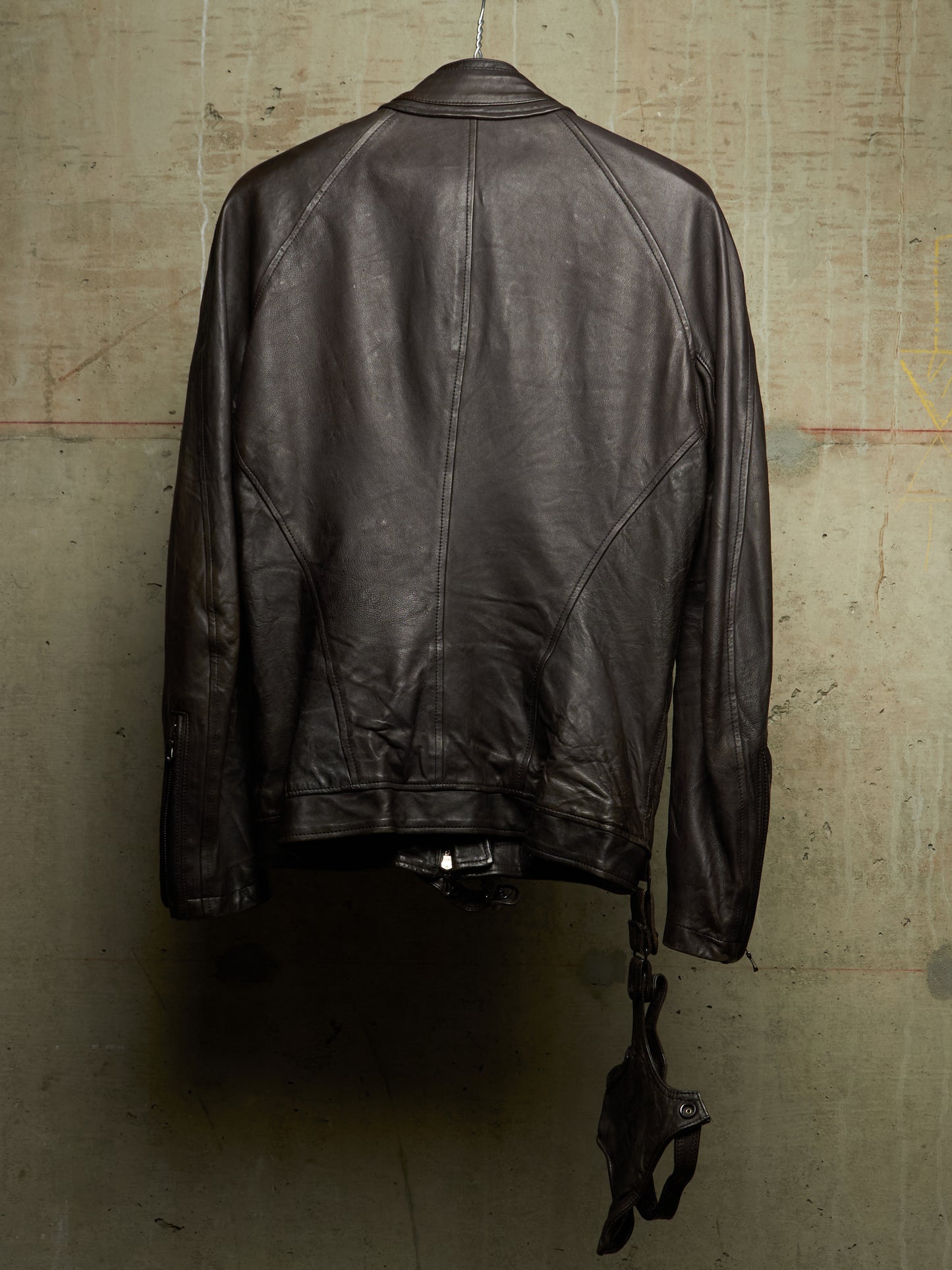 A dark brown leather jacket hanging against a textured concrete wall, shown from the back with its zipper and strap details visible.