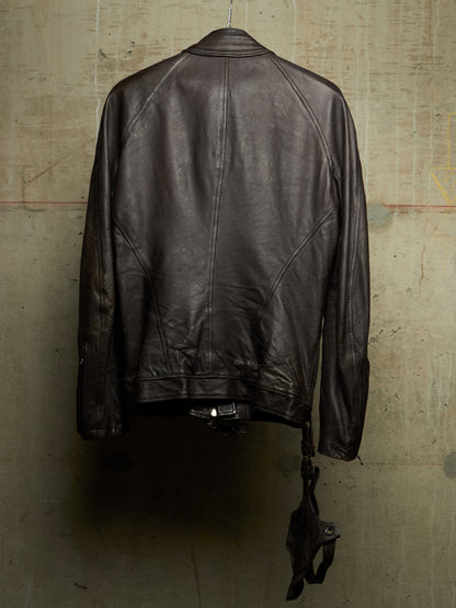 A dark brown leather jacket hanging against a textured concrete wall, shown from the back with its zipper and strap details visible.