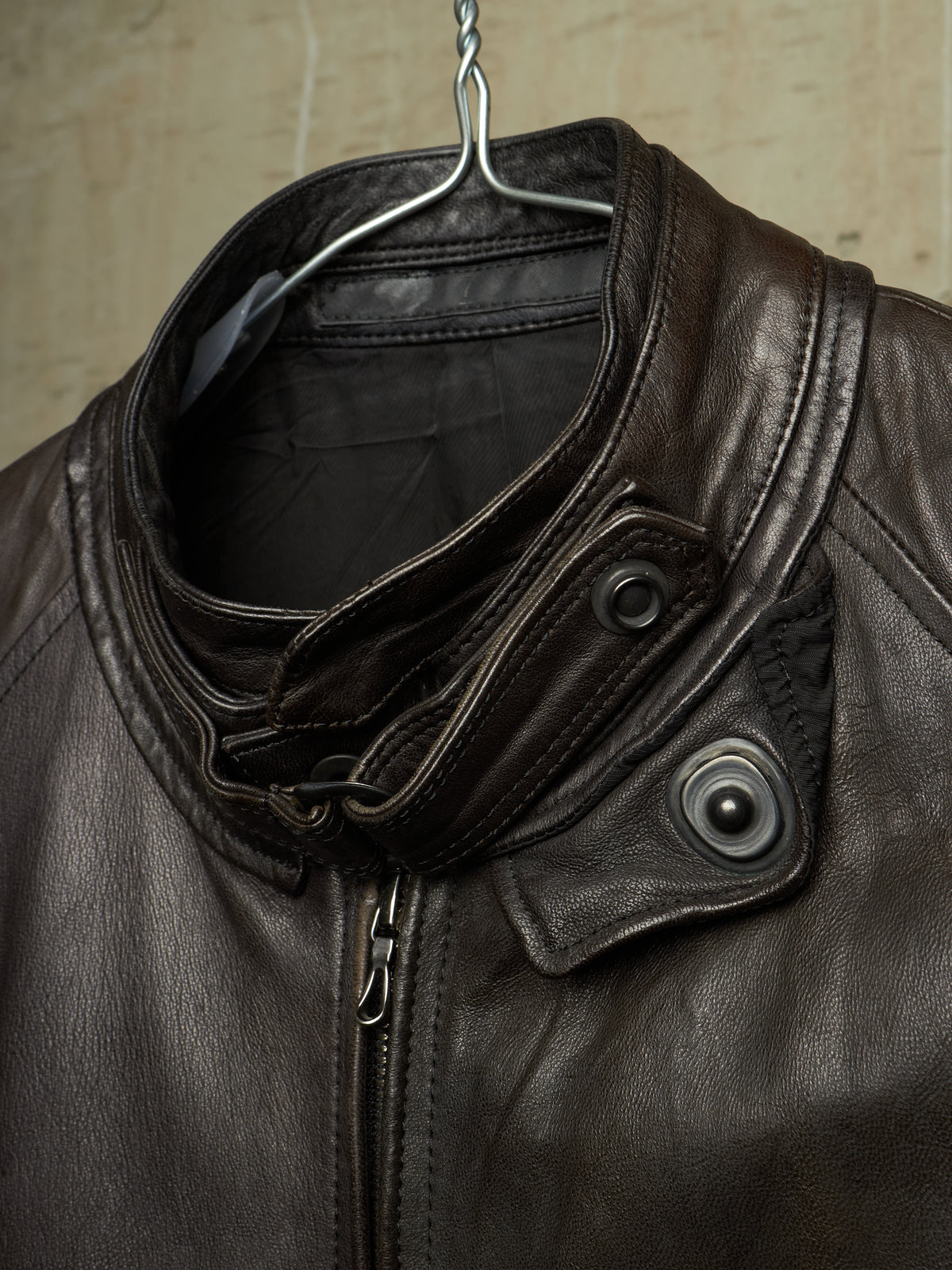Close-up of a dark brown leather jacket hanging on a metal hanger against a textured beige wall. The jacket features a high folded collar, a front zipper, and two dark snap-button flap details on the chest.