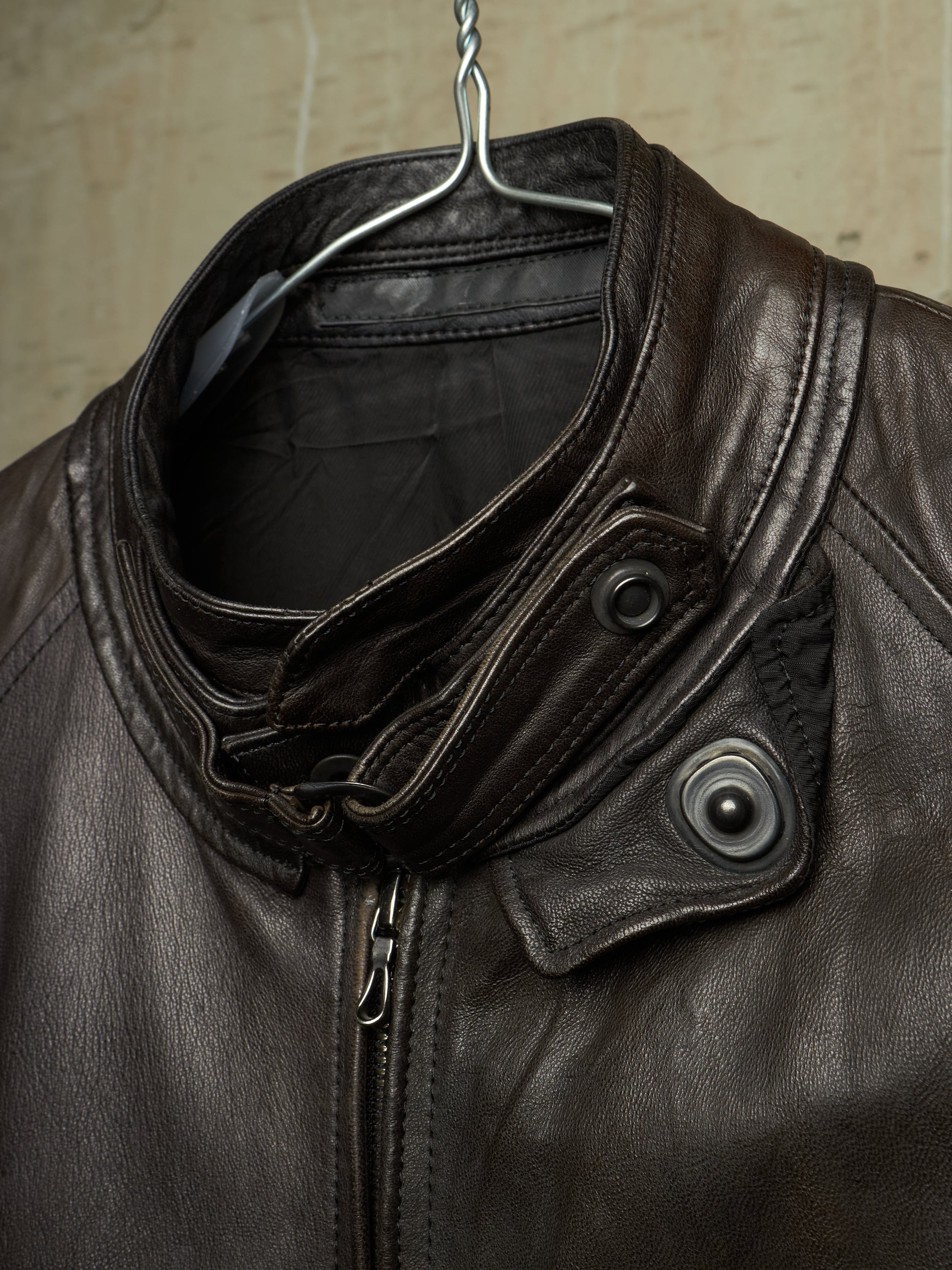Close-up of a dark brown leather jacket hanging on a metal hanger against a textured beige wall. The jacket features a high folded collar, a front zipper, and two dark snap-button flap details on the chest.