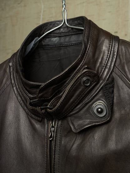 Close-up of a dark brown leather jacket hanging on a metal hanger against a textured beige wall. The jacket features a high folded collar, a front zipper, and two dark snap-button flap details on the chest.