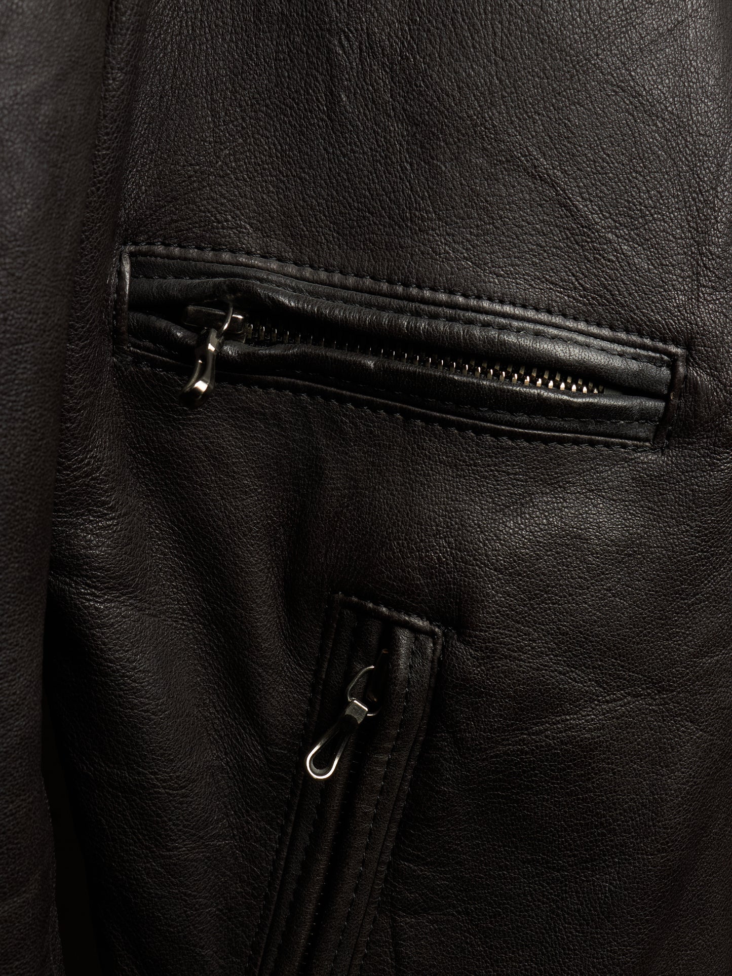 Close-up of a black leather jacket showing a zippered pocket and a vertical zip detail with silver-toned hardware.