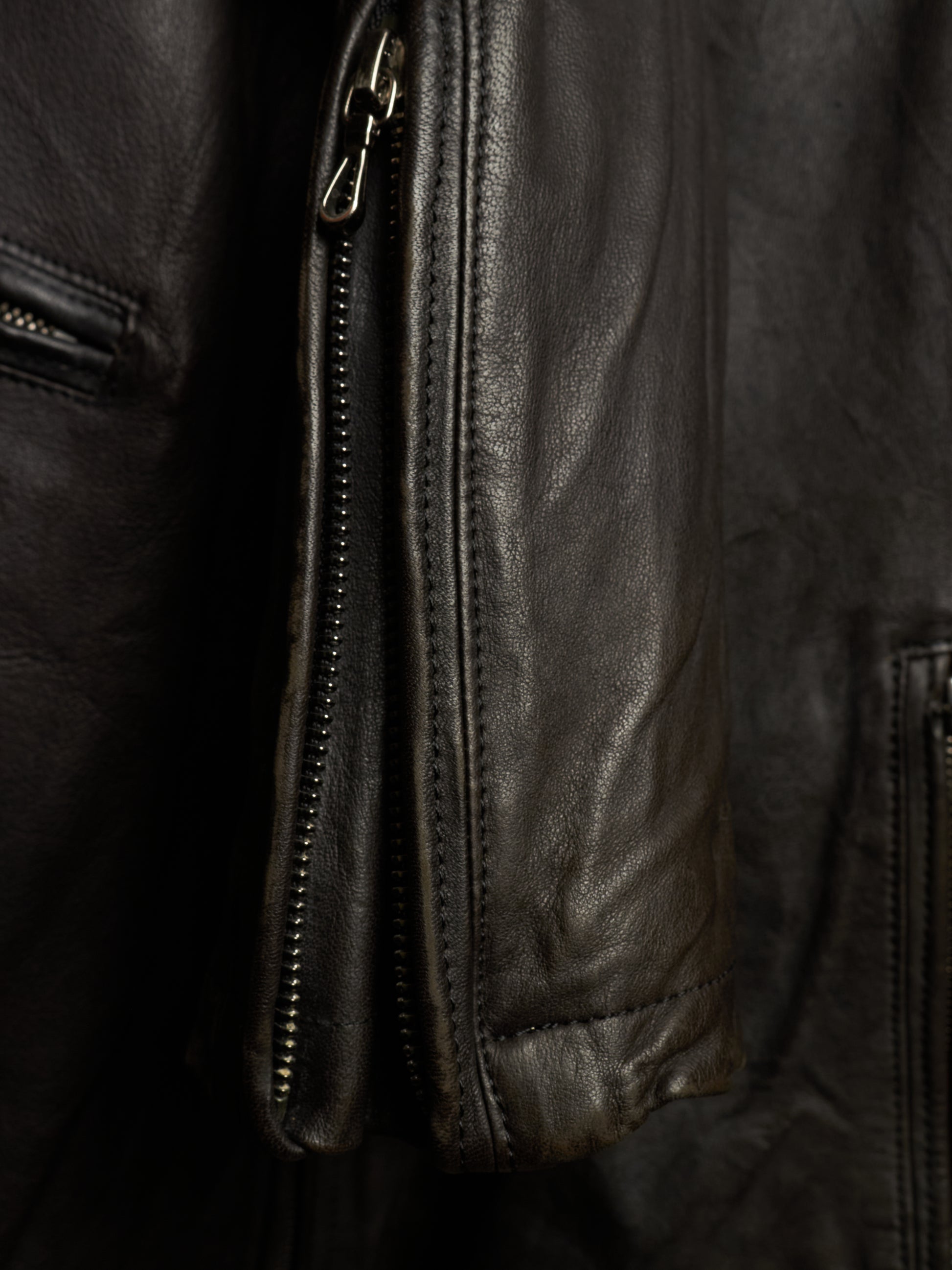 Close-up detail of a black leather jacket showing a vertical zippered pocket, stitched seams, and the textured leather surface.