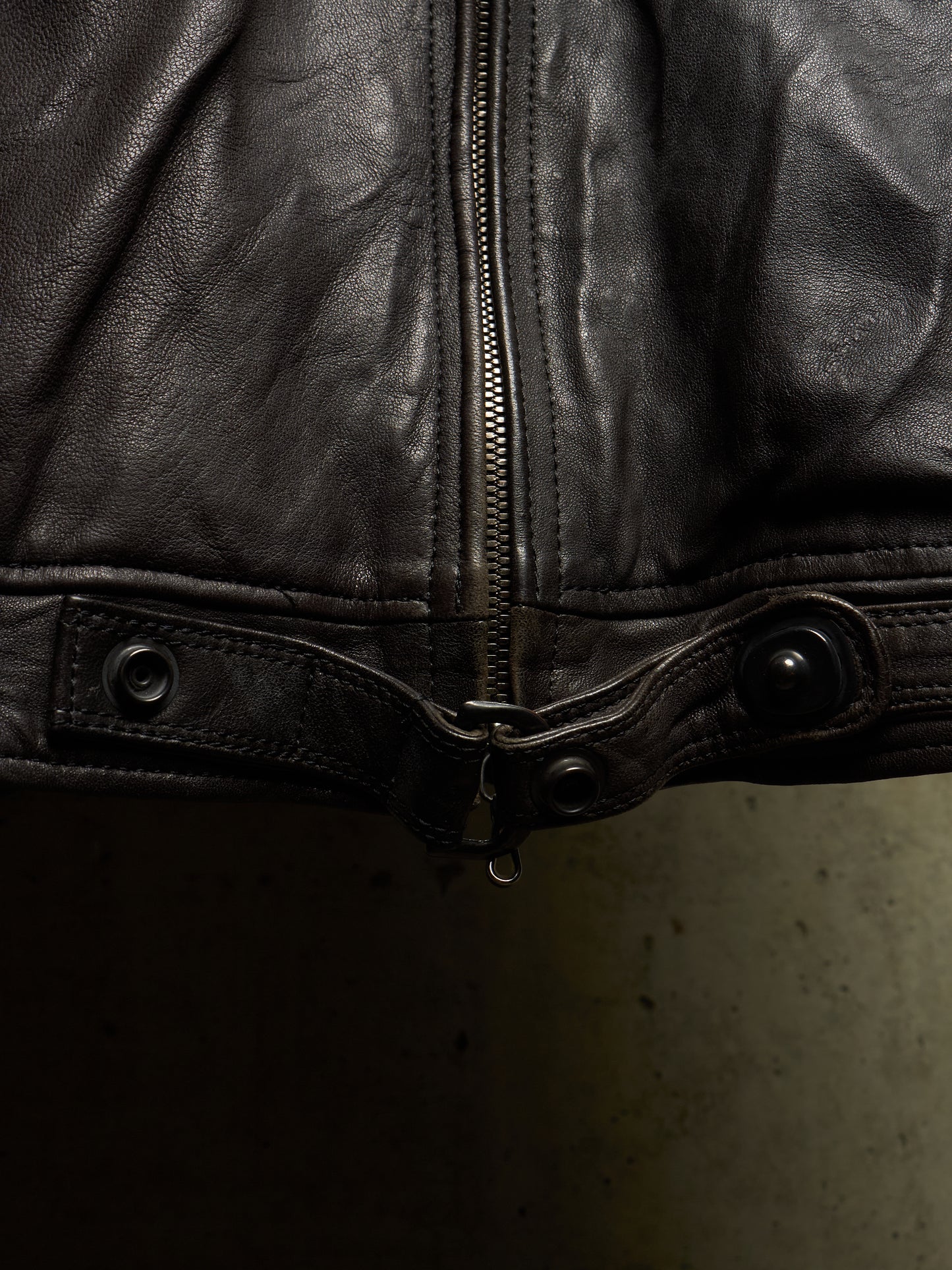 Close-up of a black leather jacket showing the front zipper, snap tab, and button details against a dark background.