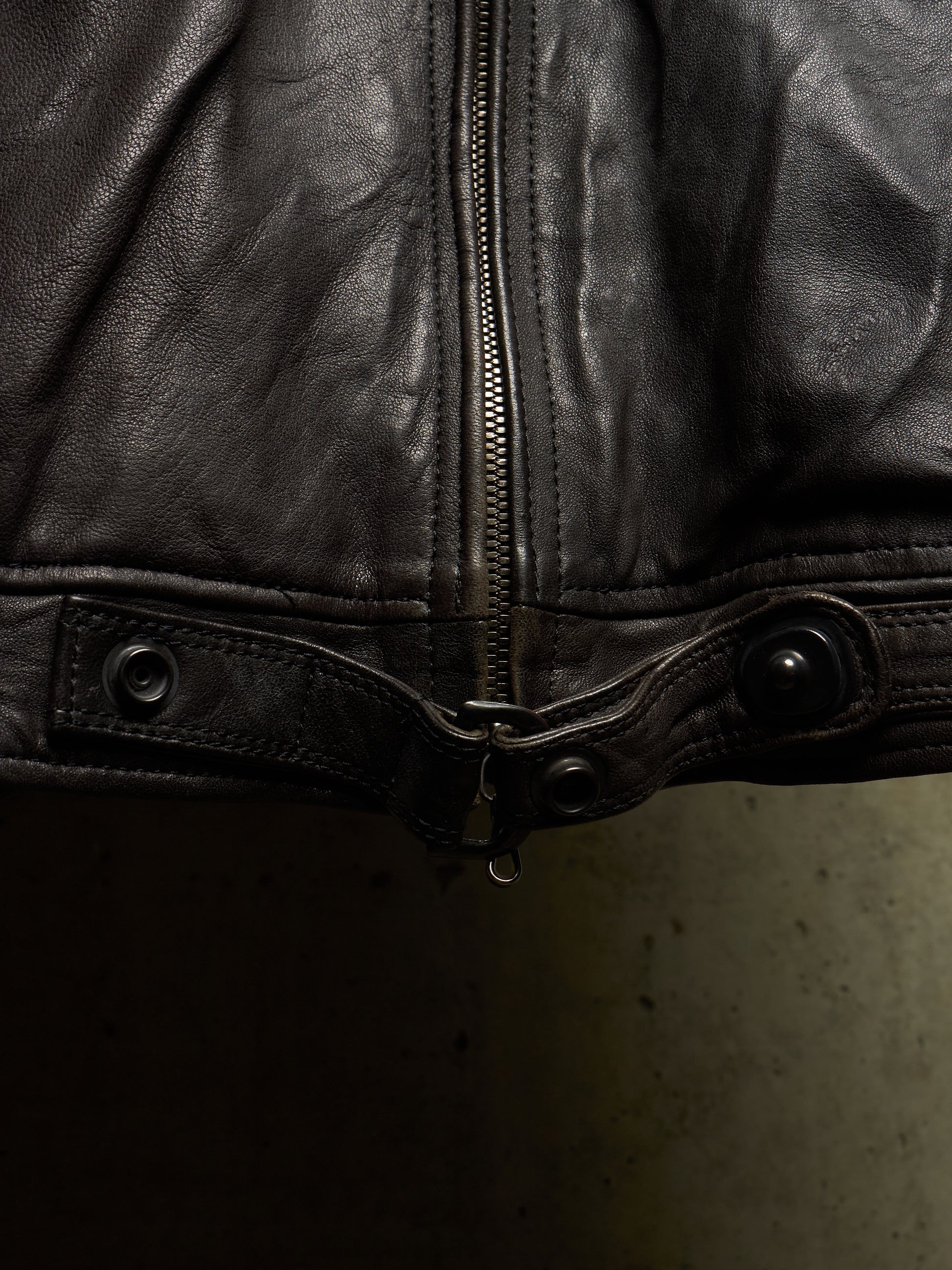 Close-up of a black leather jacket showing the front zipper, snap tab, and button details against a dark background.