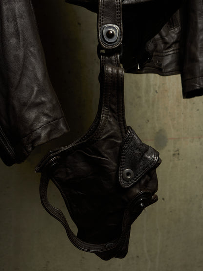A close-up of a dark brown leather motorcycle jacket hanging on a wall, showing a leather shoulder strap with a metal snap closure and a matching leather pouch or holster detail.