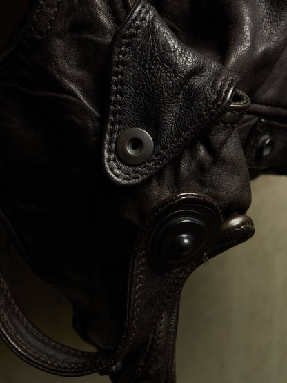 Close-up detail of a dark brown leather bag with a flap pocket, metal snap button, and textured leather strap against a muted olive background.