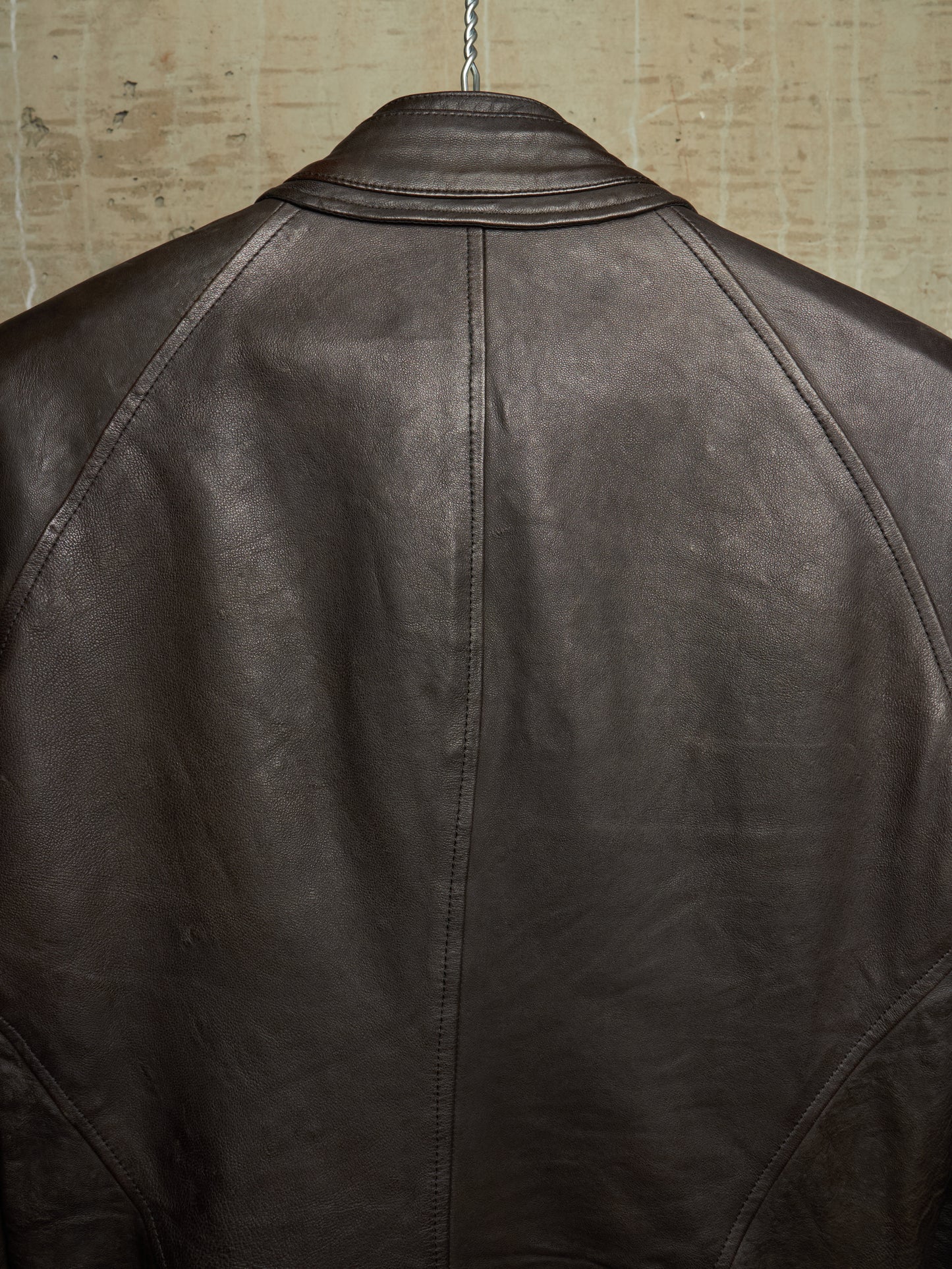Close-up back view of a dark brown leather jacket hanging against a textured beige wall.
