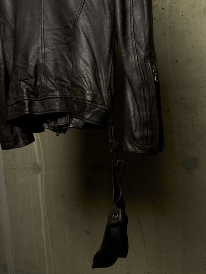 A close-up of a black leather jacket hanging against a textured concrete wall, showing the sleeve with a zipper pocket and a dangling black strap or pouch.
