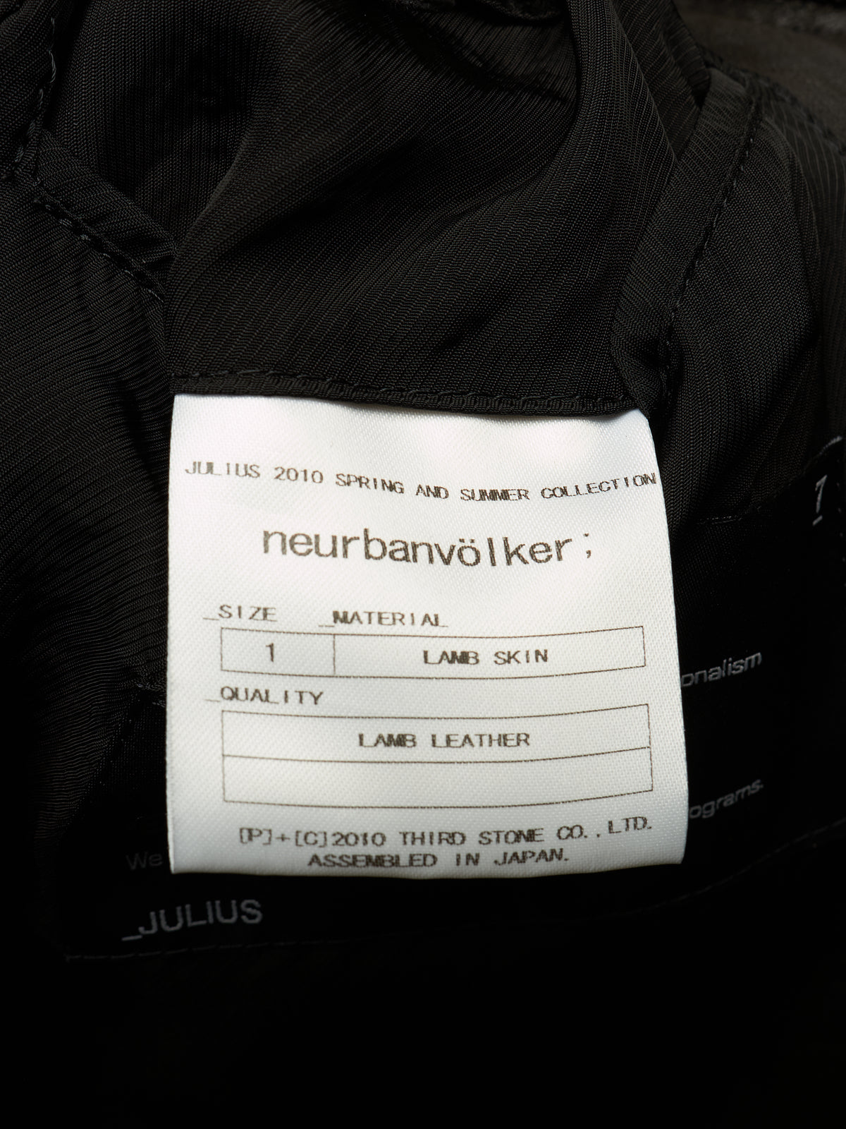 Close-up of a white fabric product label sewn onto a black garment, showing the brand name 'neurbanvölker' and text for Julius 2010 Spring and Summer Collection, size 1, lamb skin, and lamb leather.