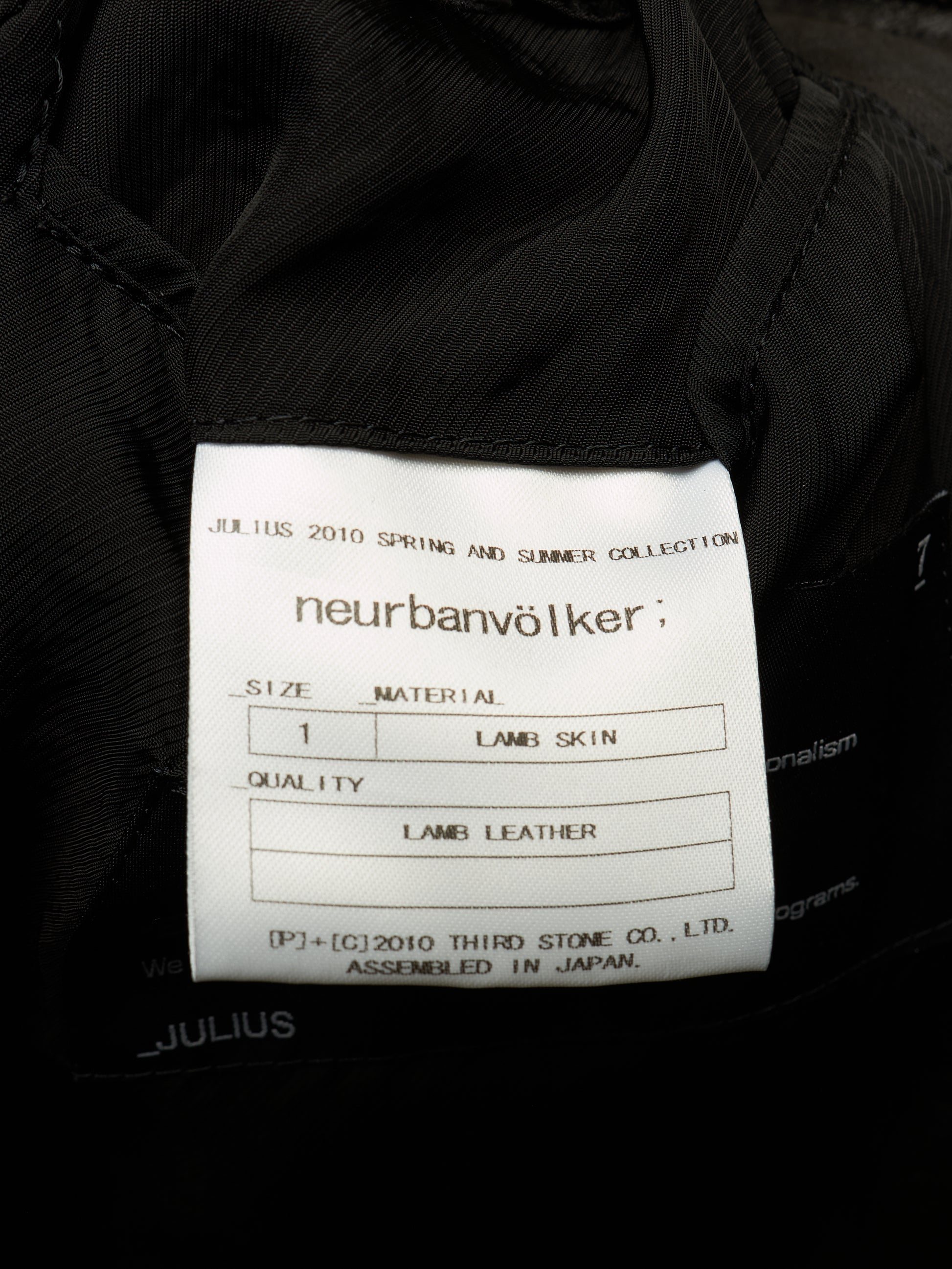 Close-up of a white fabric product label sewn onto a black garment, showing the brand name 'neurbanvölker' and text for Julius 2010 Spring and Summer Collection, size 1, lamb skin, and lamb leather.
