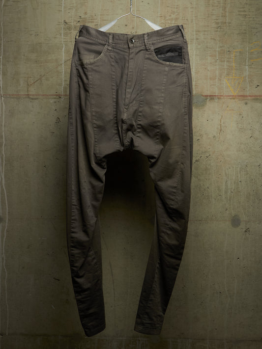 A pair of dark brown, relaxed-fit pants with a unique draped, baggy silhouette, hanging from a white plastic hanger against a textured concrete wall.