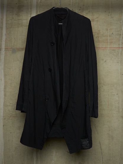 A black long-sleeve open-front coat with a high collar and three front buttons, hanging against a textured concrete wall.