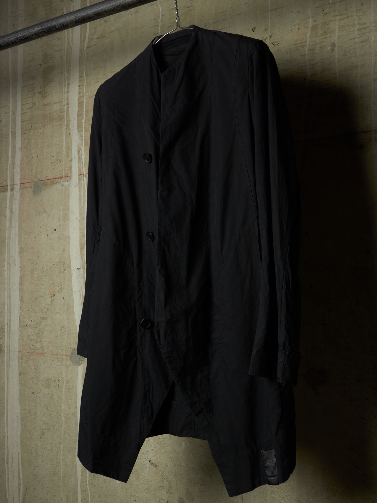 A black long-sleeve button-front jacket or overshirt hanging on a hanger against a textured concrete wall.