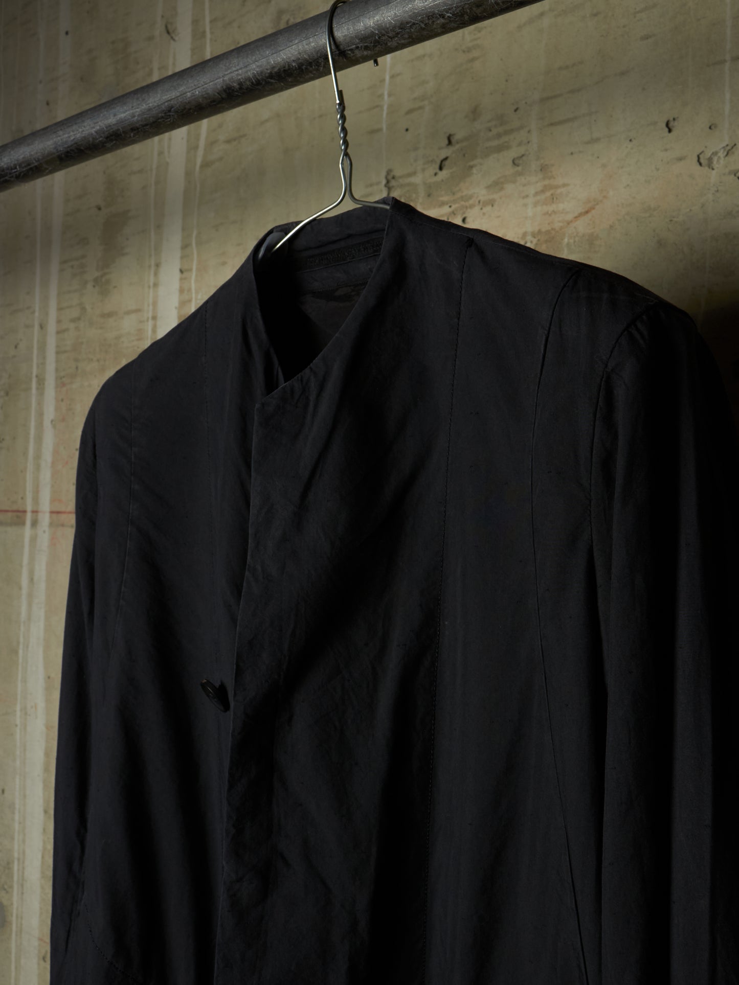 A black long-sleeve shirt with a high collar and front button placket, hanging on a metal hanger from a dark industrial rod against a textured concrete wall.