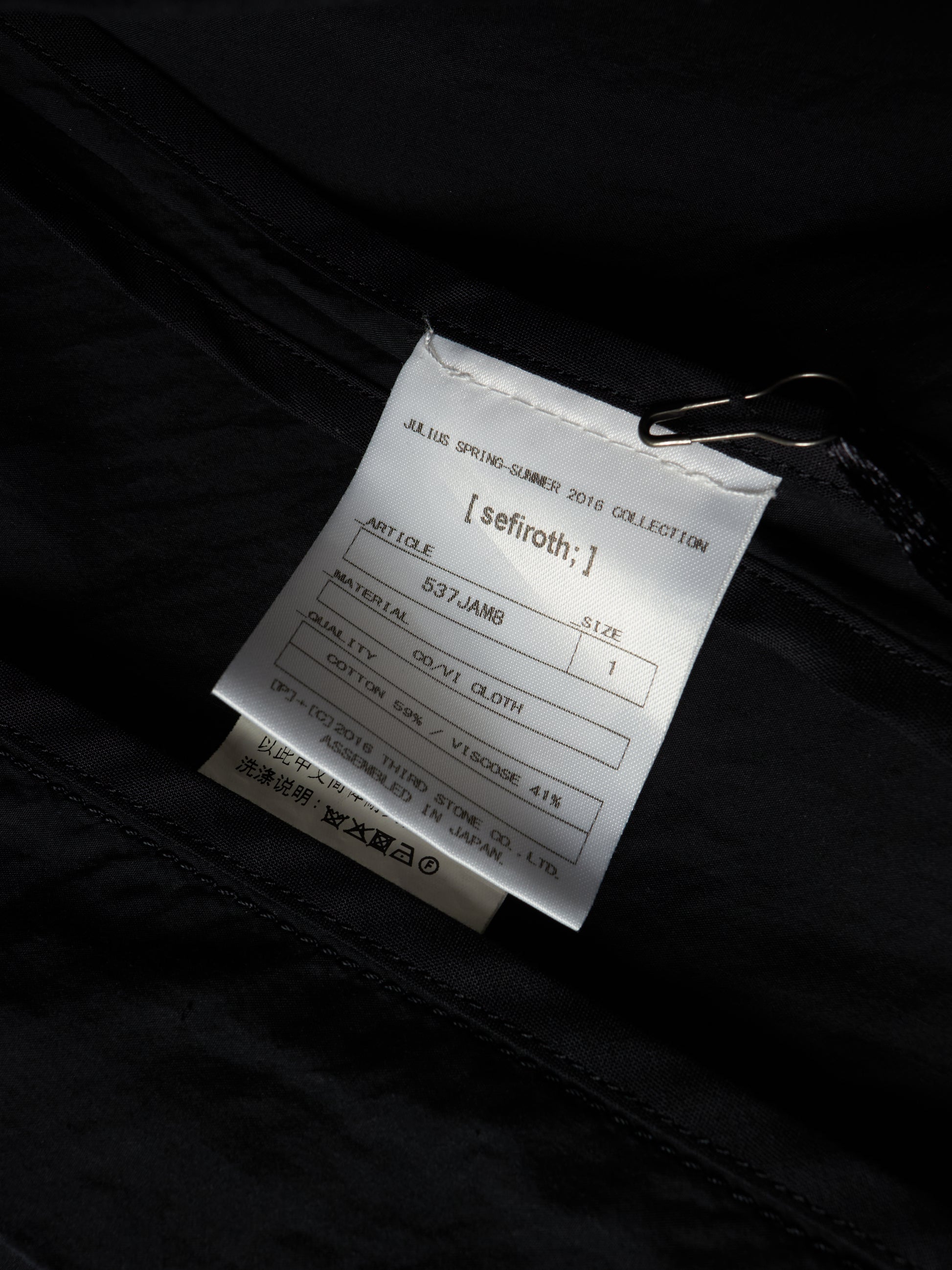 A close-up of a white product tag attached to a black garment with a metal clip, showing printed text including the brand name 'seifroth;'.