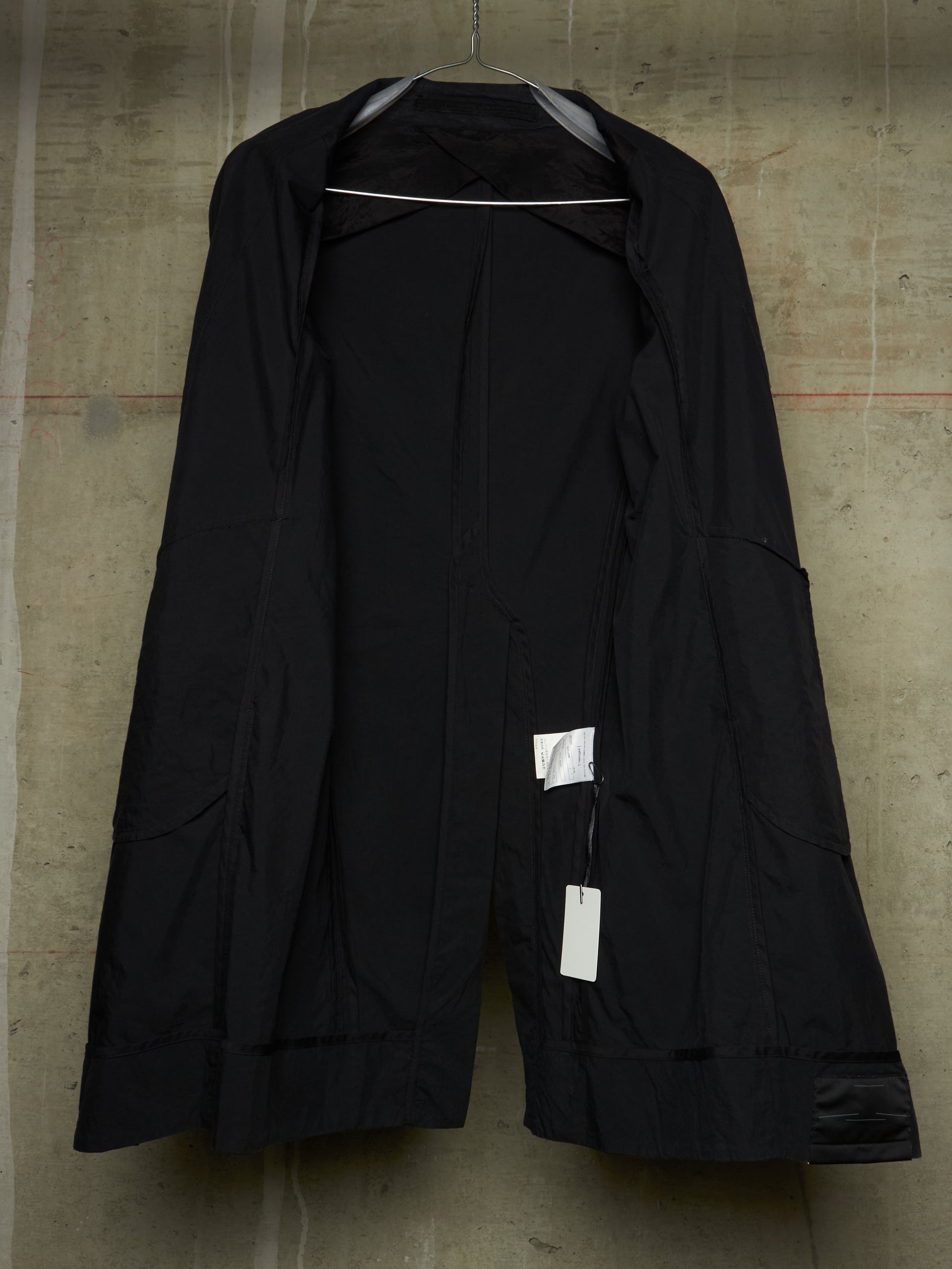 A black open-front jacket or coat hanging on a hanger against a textured concrete wall, with white retail tags attached near the lower front.