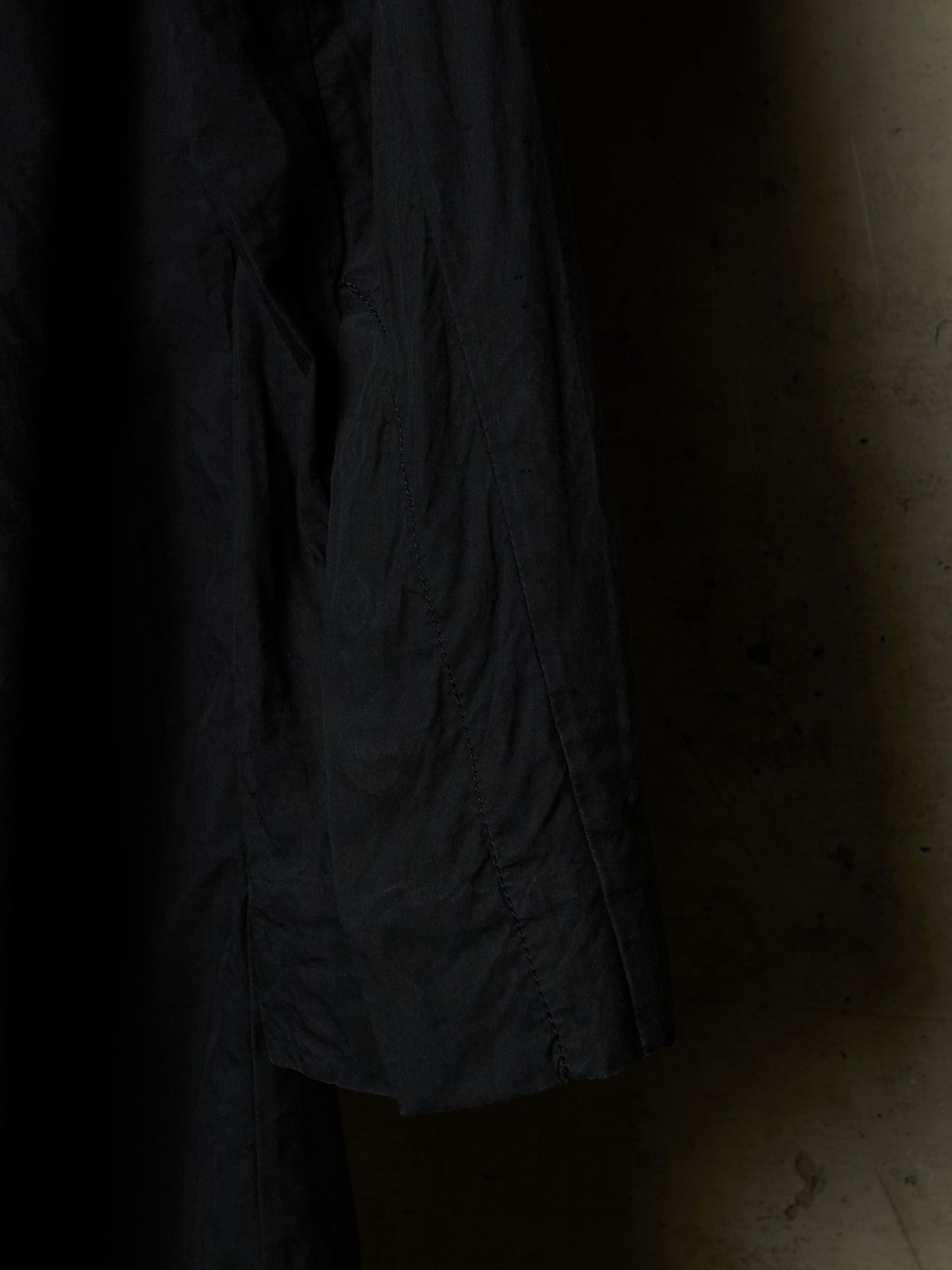 A close-up of a dark charcoal gray or black textured fabric, likely a jacket or coat sleeve, hanging against a dimly lit, warm-toned concrete wall.