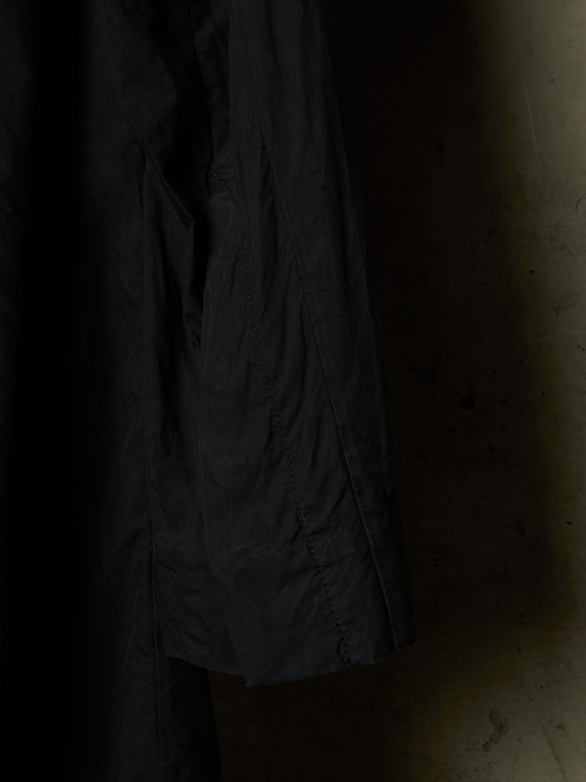A close-up of a dark charcoal gray or black textured fabric, likely a jacket or coat sleeve, hanging against a dimly lit, warm-toned concrete wall.