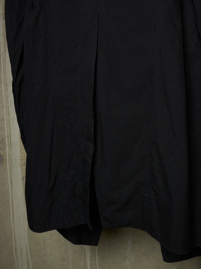 A close-up of a black garment, likely a skirt or dress, showing the fabric texture and a vertical seam, hanging against a textured concrete wall.