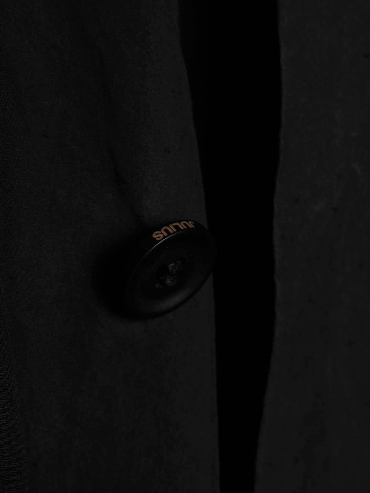 Close-up of a dark charcoal wool-blend coat with a single black button featuring gold 'silk' branding, set against a dark background.