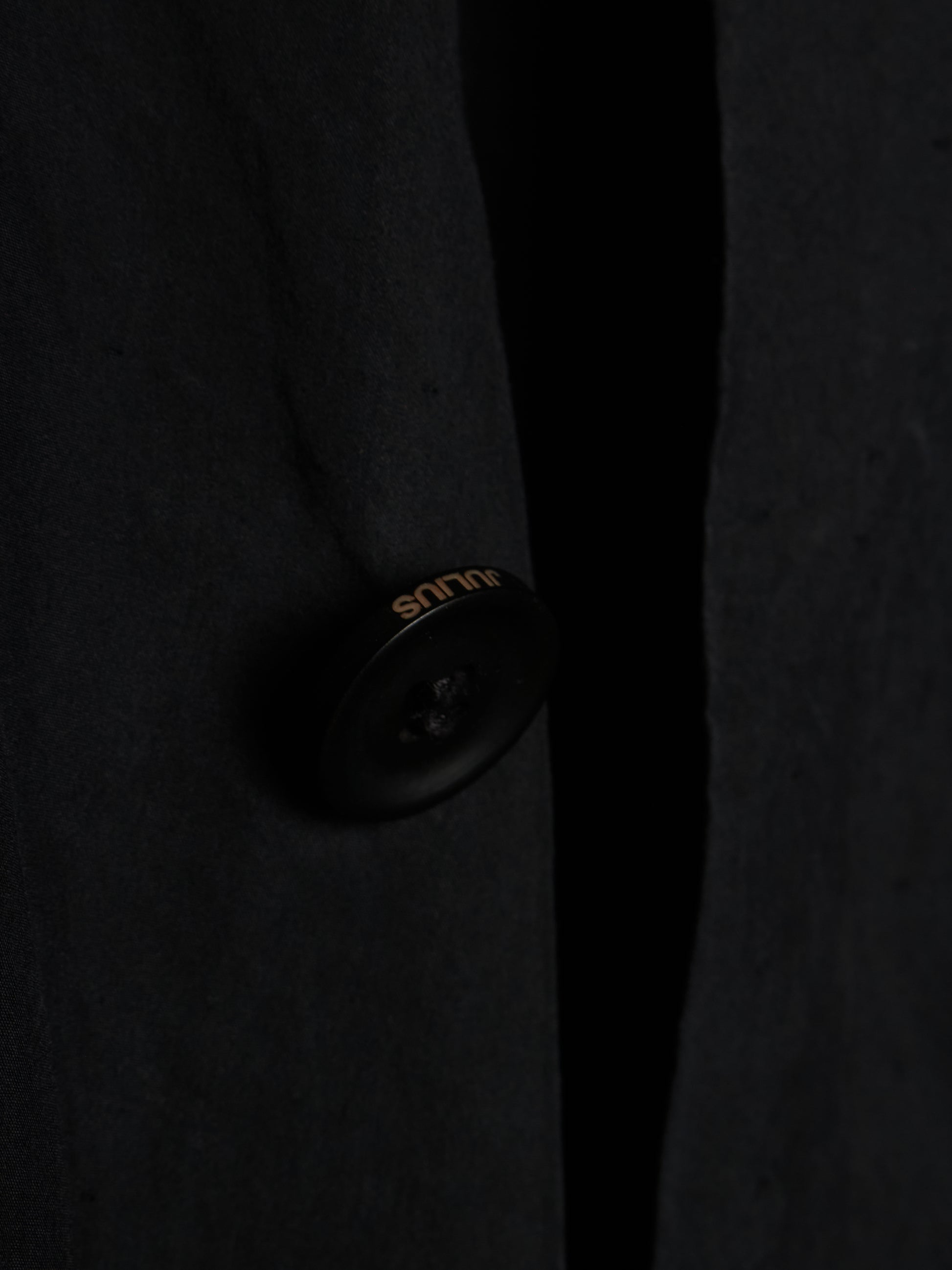 Close-up of a dark charcoal wool-blend coat with a single black button featuring gold 'silk' branding, set against a dark background.