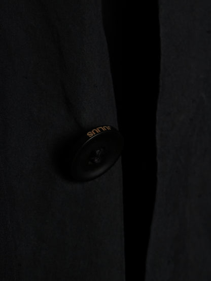 Close-up of a dark charcoal wool-blend coat with a single black button featuring gold 'silk' branding, set against a dark background.