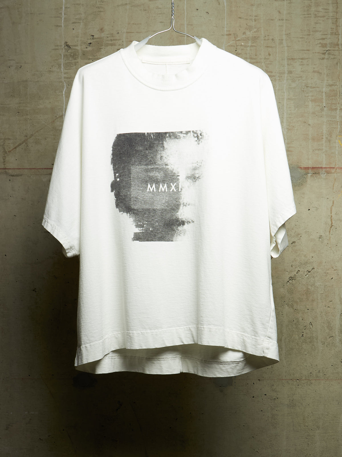 A white oversized short-sleeve t-shirt hanging on a wire hanger against a textured concrete wall. The shirt features a large rectangular black and gray graphic print on the chest with the Roman numerals 'MMXI' printed in white.