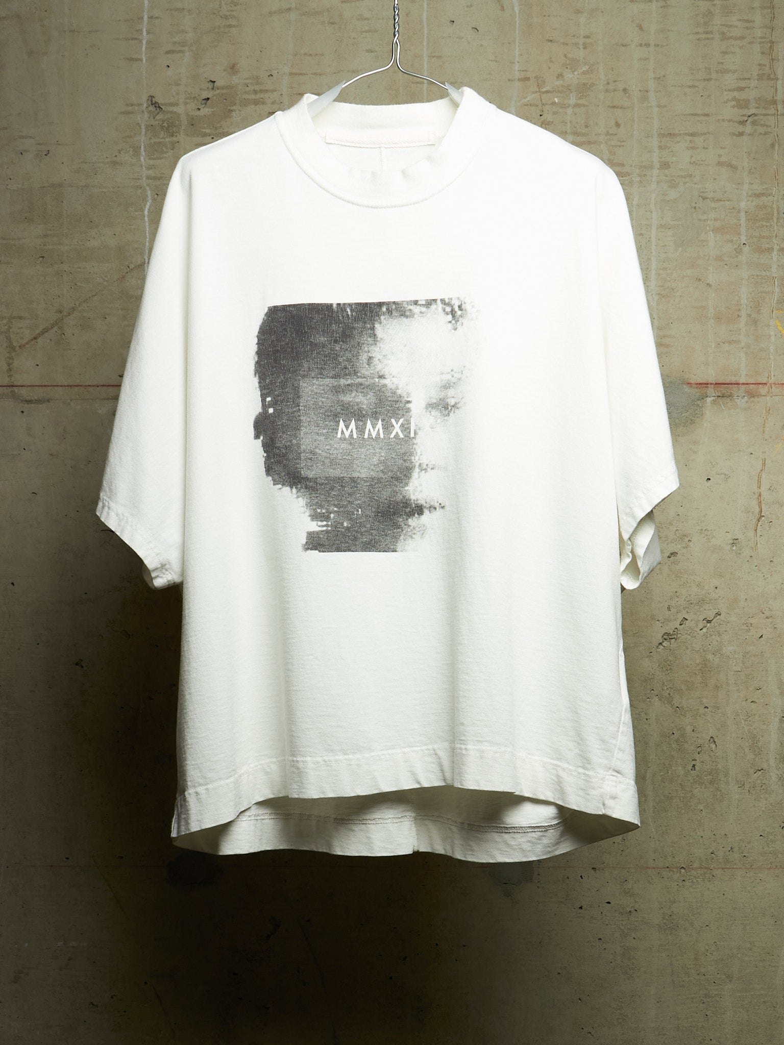 A white oversized short-sleeve t-shirt hanging on a wire hanger against a textured concrete wall. The shirt features a large rectangular black and gray graphic print on the chest with the Roman numerals 'MMXI' printed in white.