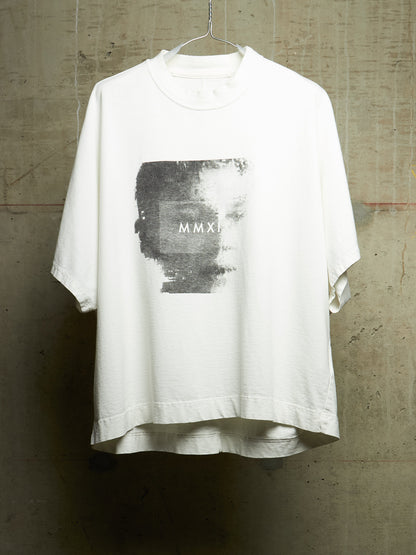 A white oversized short-sleeve t-shirt hanging on a wire hanger against a textured concrete wall. The shirt features a large rectangular black and gray graphic print on the chest with the Roman numerals 'MMXI' printed in white.
