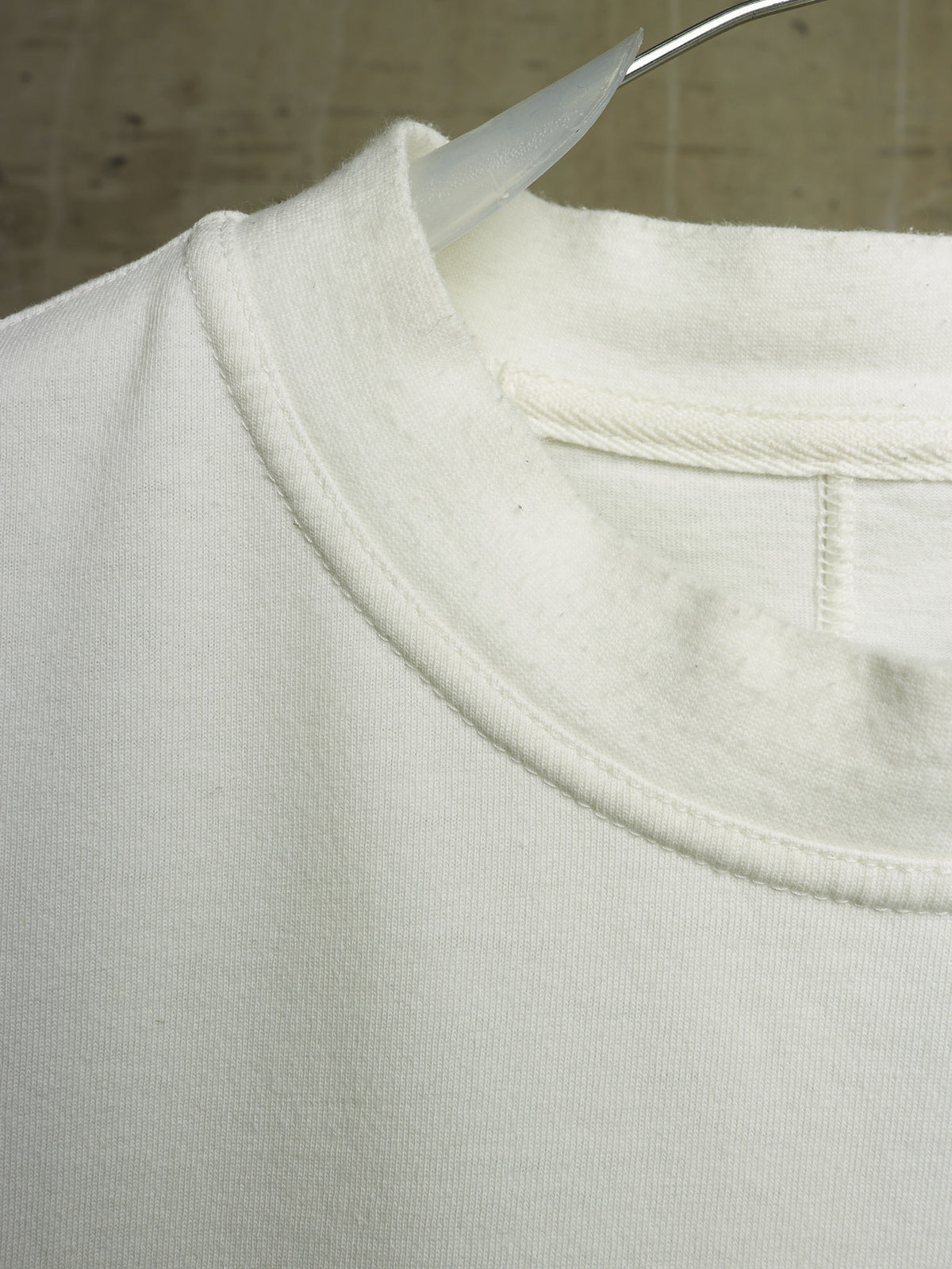 Close-up of a white crewneck sweatshirt hanging on a clear plastic hanger against a neutral textured background.