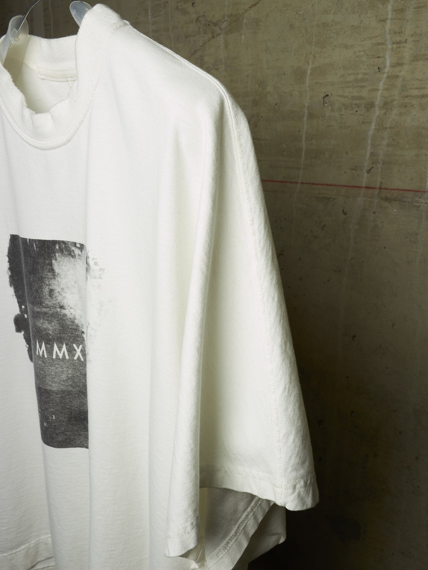 A white oversized short-sleeve t-shirt hanging on a clear plastic hanger against a textured concrete wall. The shirt features a black rectangular graphic print on the front with the Roman numerals 'MMX'.