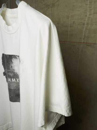 A white oversized short-sleeve t-shirt hanging on a clear plastic hanger against a textured concrete wall. The shirt features a black rectangular graphic print on the front with the Roman numerals 'MMX'.