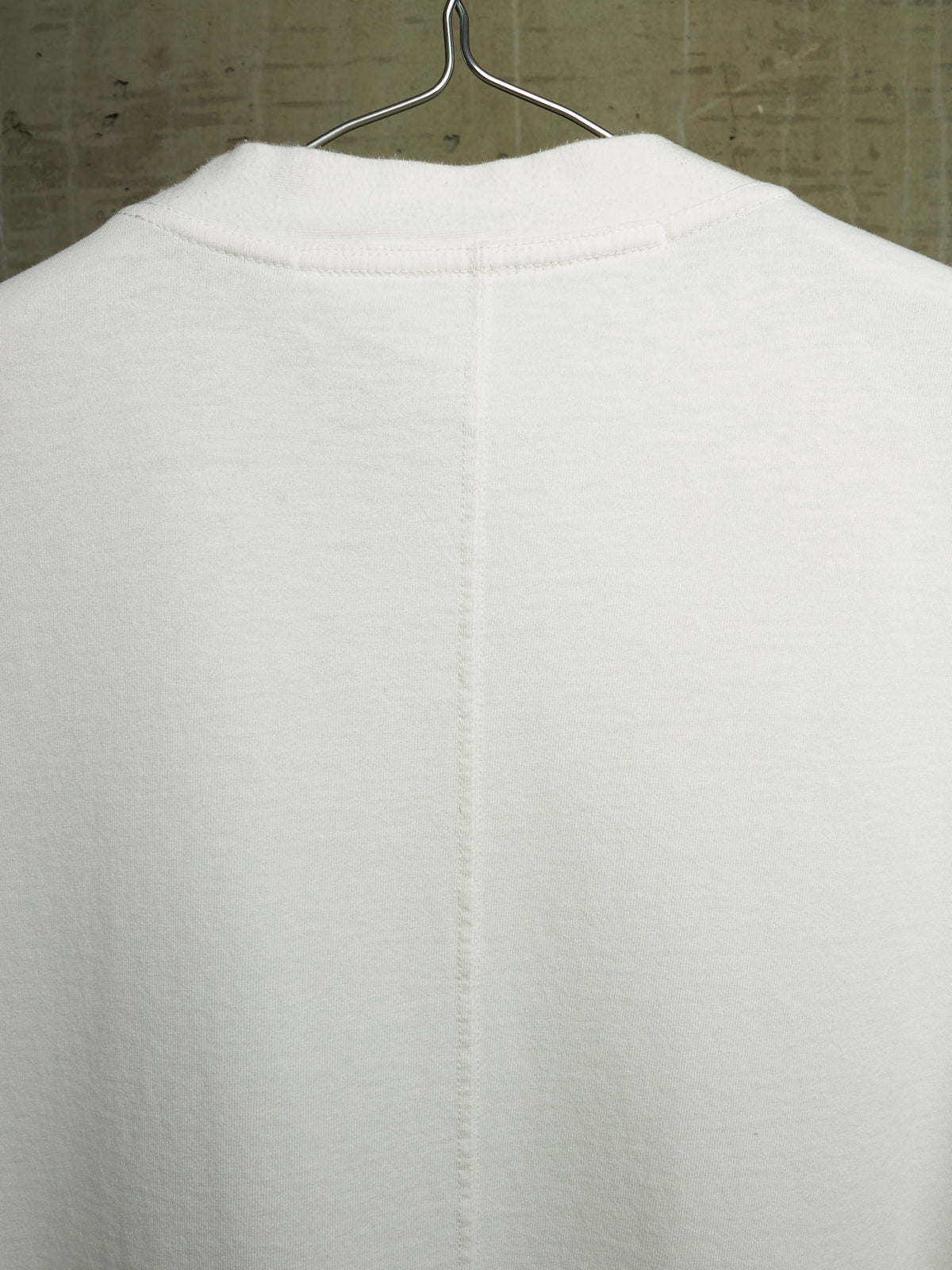 A close-up of the back of a white knit garment hanging on a metal hanger against a textured gray wall.