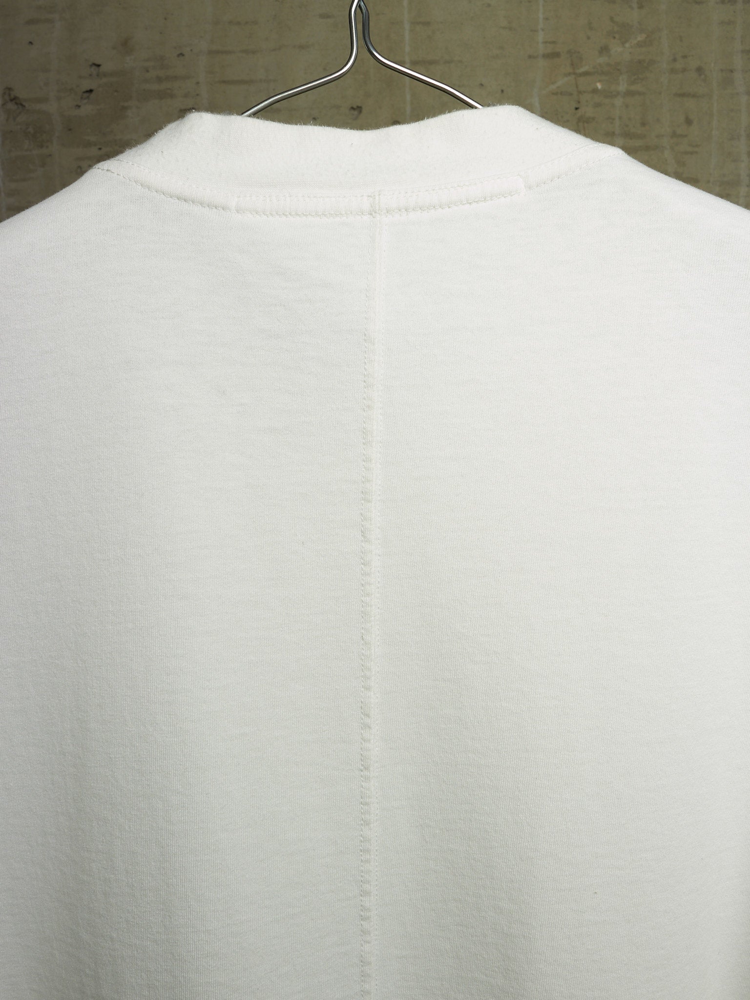 A close-up of the back of a white knit garment hanging on a metal hanger against a textured gray wall.