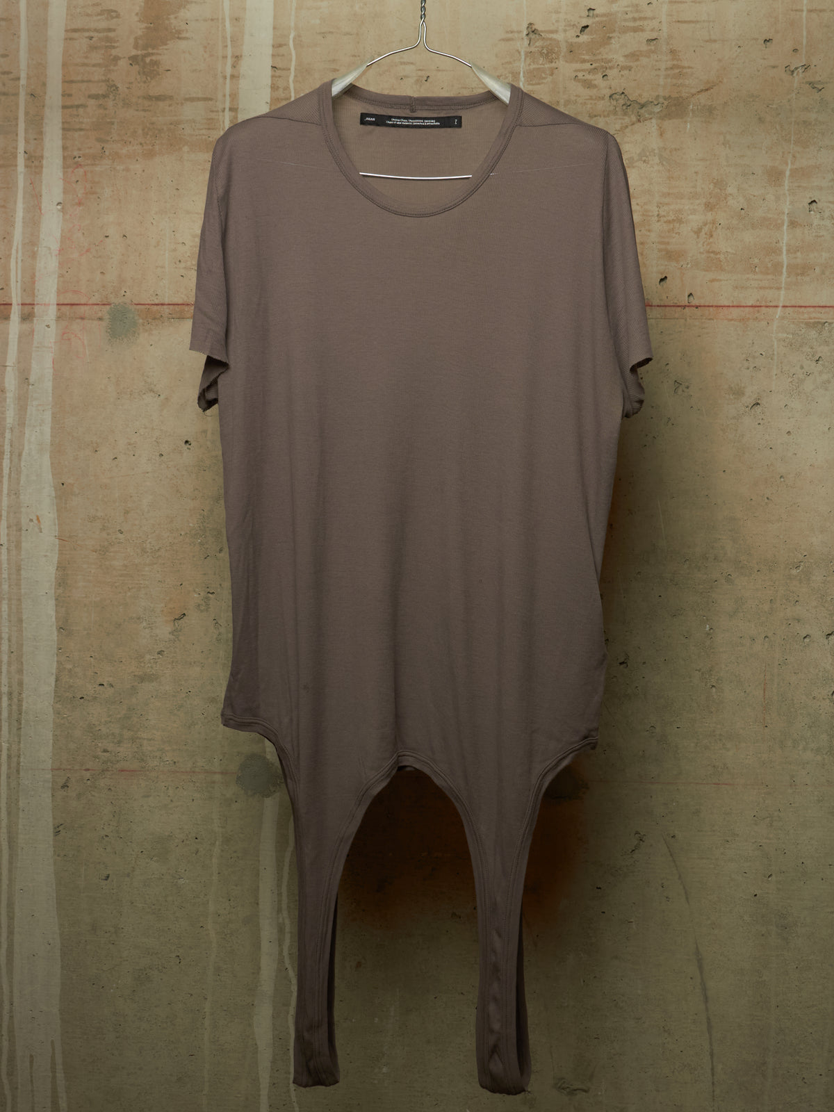 A taupe-brown short-sleeve tunic top with a split leg design, hanging on a white hanger against a textured concrete wall.