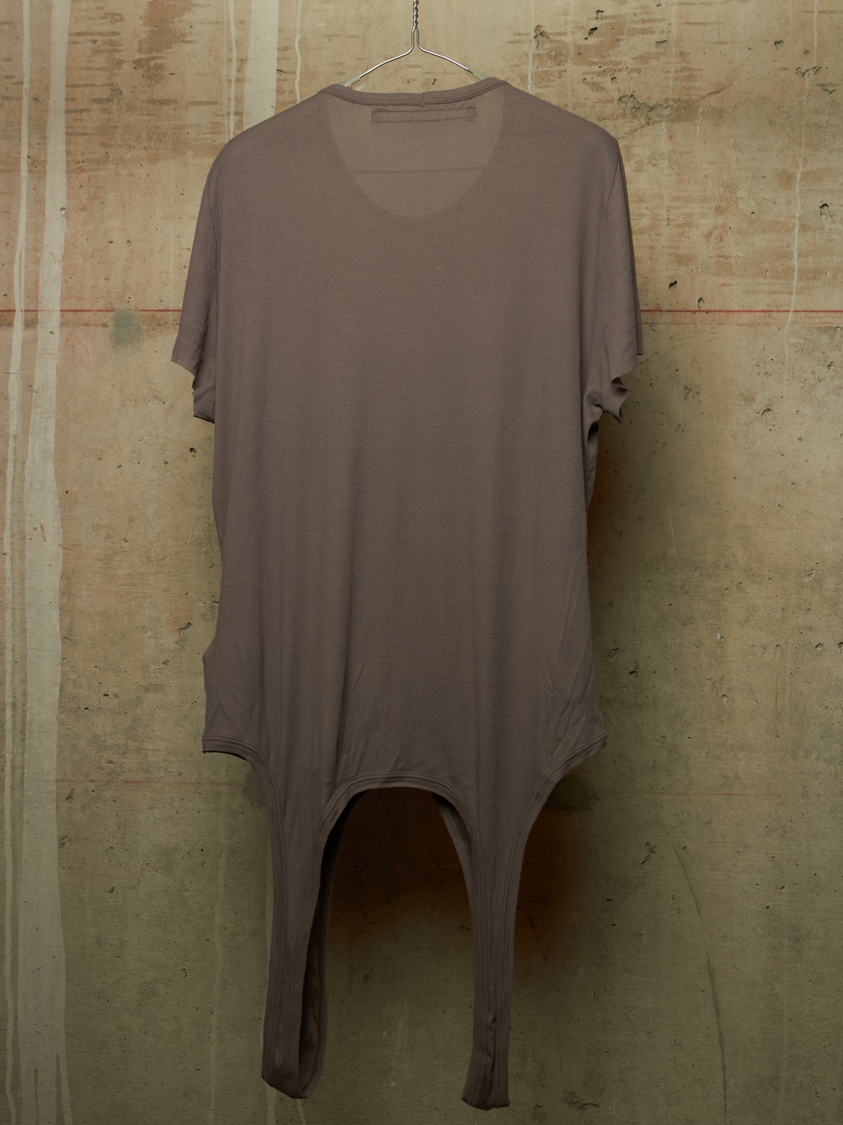 A loose-fitting, short-sleeve tunic dress in a muted taupe-brown color, hanging on a wire hanger against a textured concrete wall.