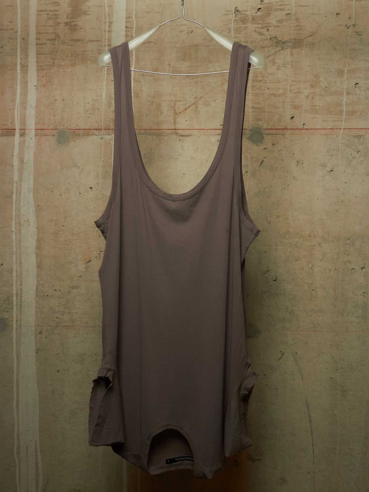A sleeveless scoop-neck tank top in a muted taupe-brown color, hanging on a white wire hanger against a textured concrete wall.